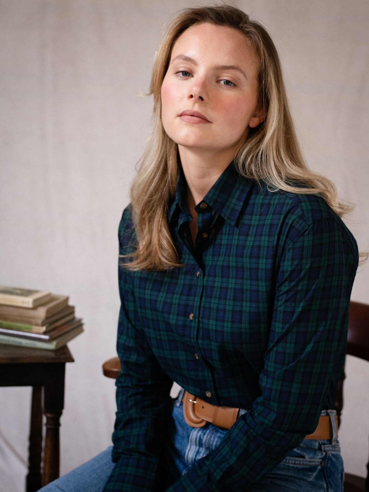 A woman with long blonde hair wears a Blackwatch Cotton Tartan Shirt by Campbell's of Beauly and jeans, sitting indoors near a small table with stacked books, looking calmly at the camera.