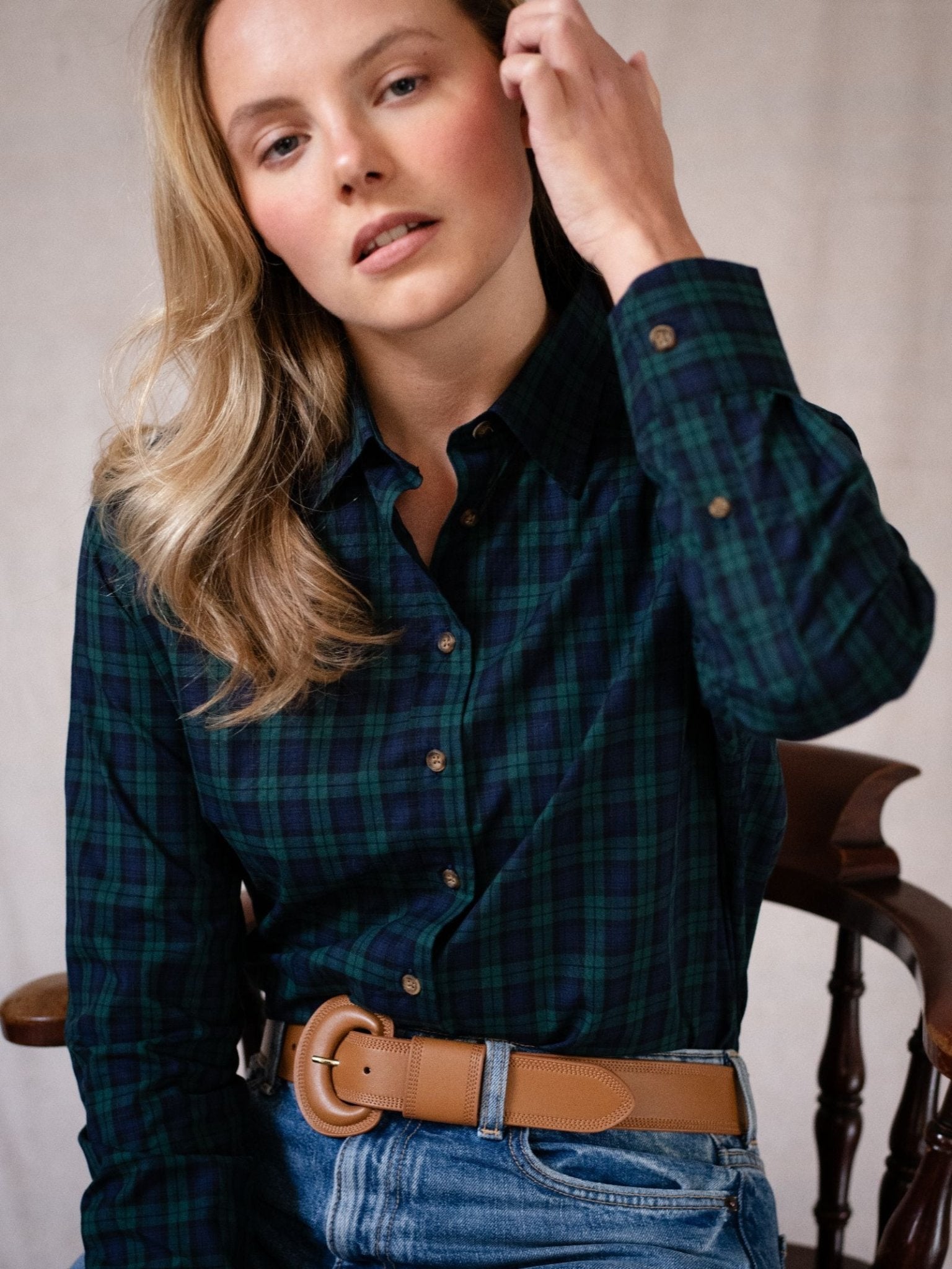 A woman with long blonde hair wears the Campbell's of Beauly Cotton Tartan Shirt, blue jeans, and a tan belt. Seated on a wooden chair, she touches her hair while looking at the camera.