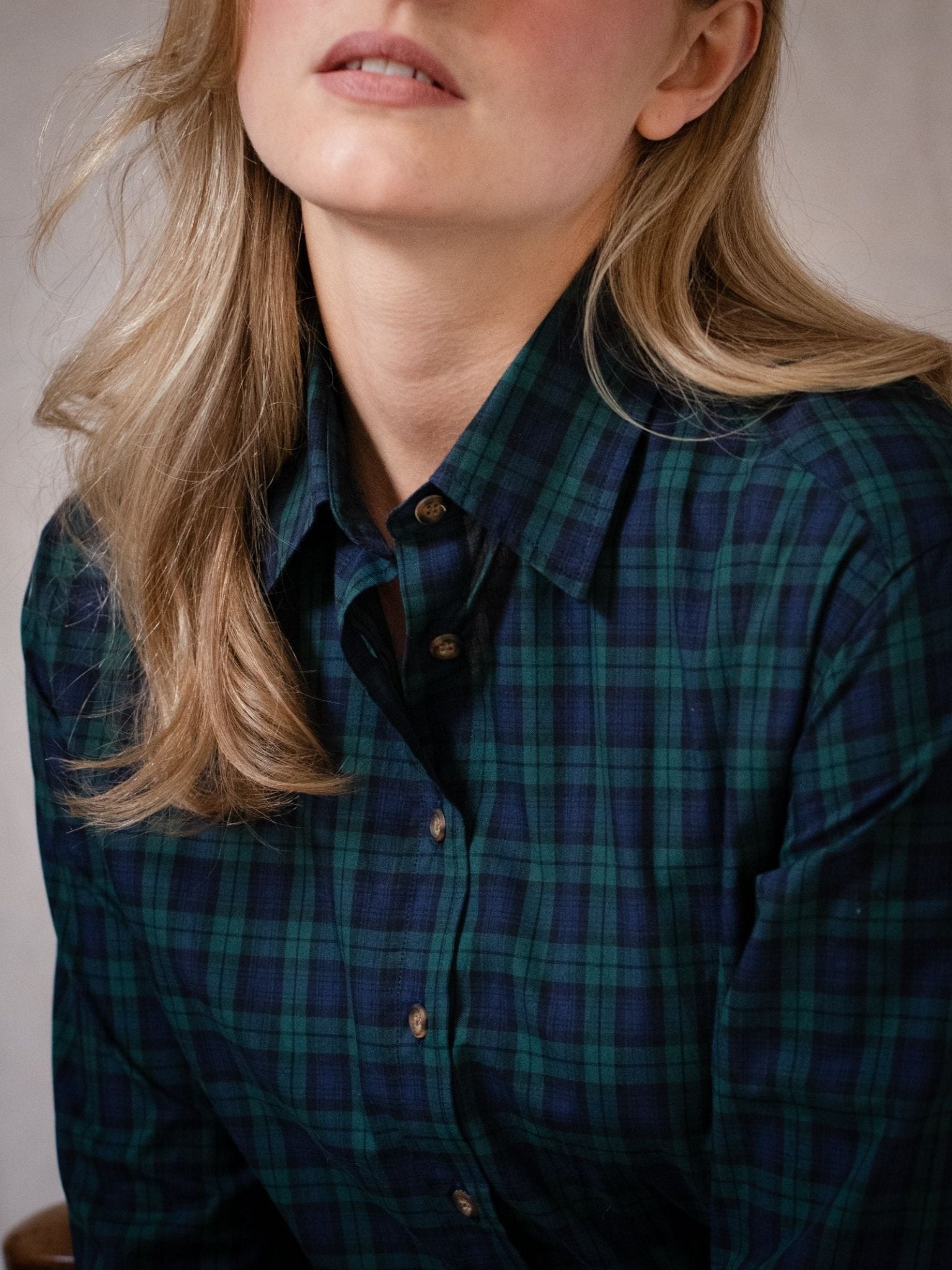 A close-up shows a woman with long blonde hair wearing the Campbell's of Beauly Cotton Tartan Shirt in dark green and navy blue. Her face is partially visible as she looks slightly upwards.