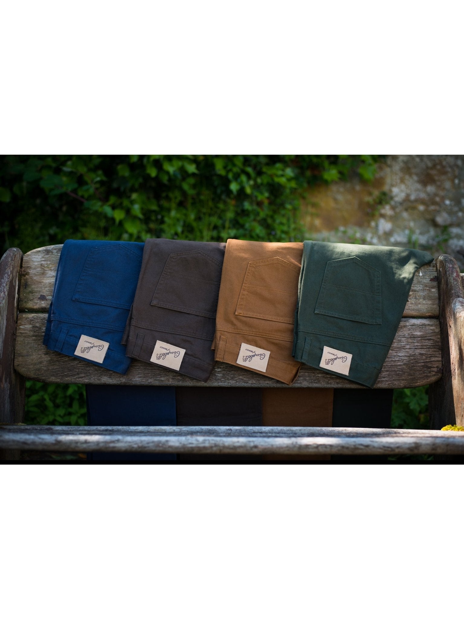 Four pairs of Neutral Cotton Jeans by Campbell's of Beauly in blue, brown, tan, and green, each with a straight leg and classic five pocket design, are neatly folded on an outdoor wooden bench with greenery behind.