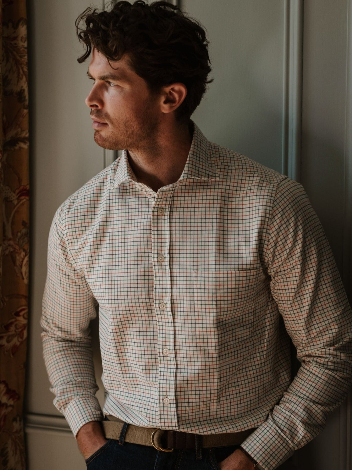 A man with curly brown hair, wearing the Campbell’s of Beauly Windowpane Shirt in a red, blue, and green check pattern and a beige belt, stands indoors near a window with floral curtains, looking to the side.