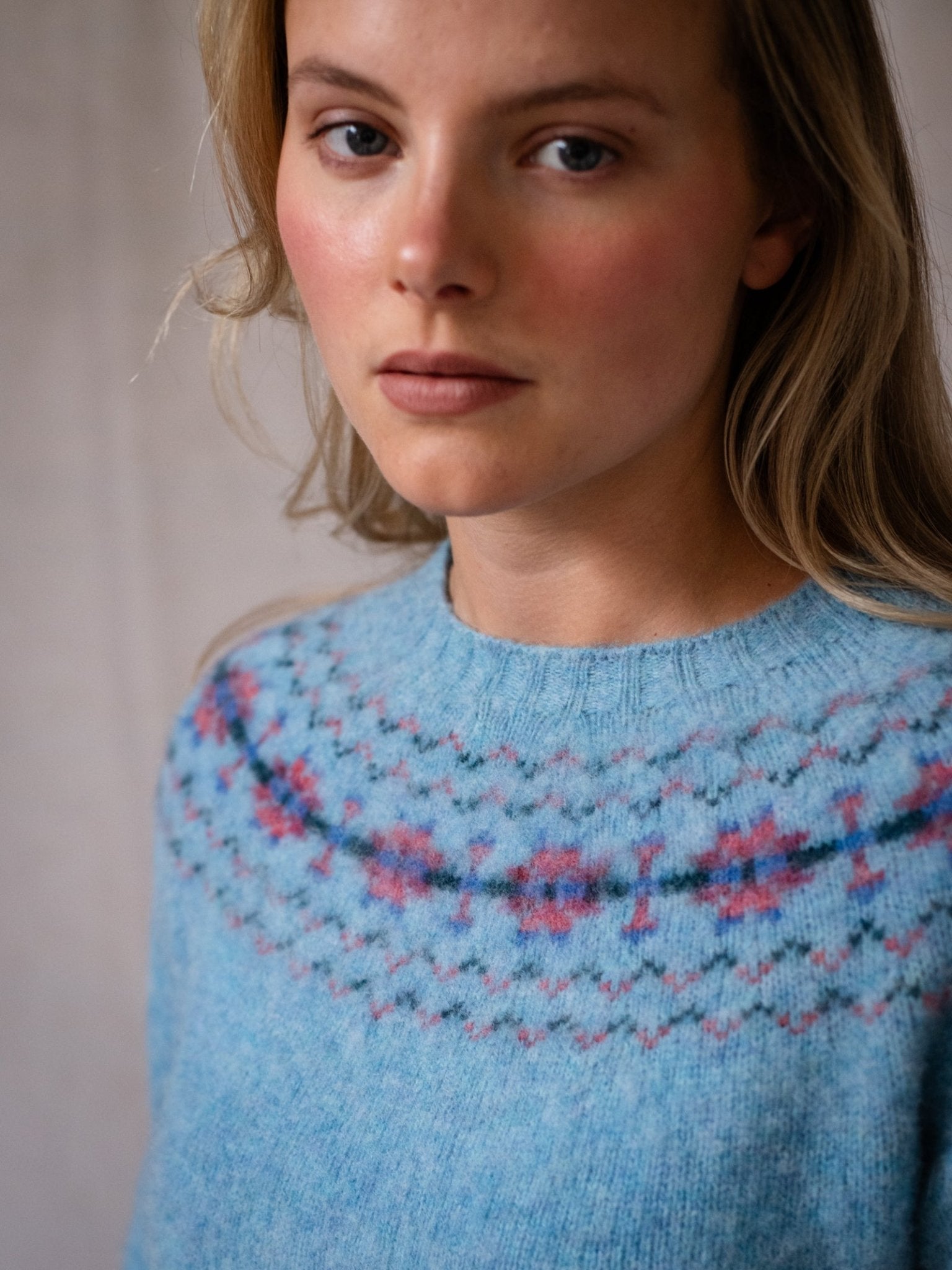 A woman with light skin and long blonde hair wears the Campbell's of Beauly Shaggy Cropped Fairisle Crew Jumper in light blue, featuring a pink and blue geometric pattern around the neckline, gazing at the camera with a neutral expression.