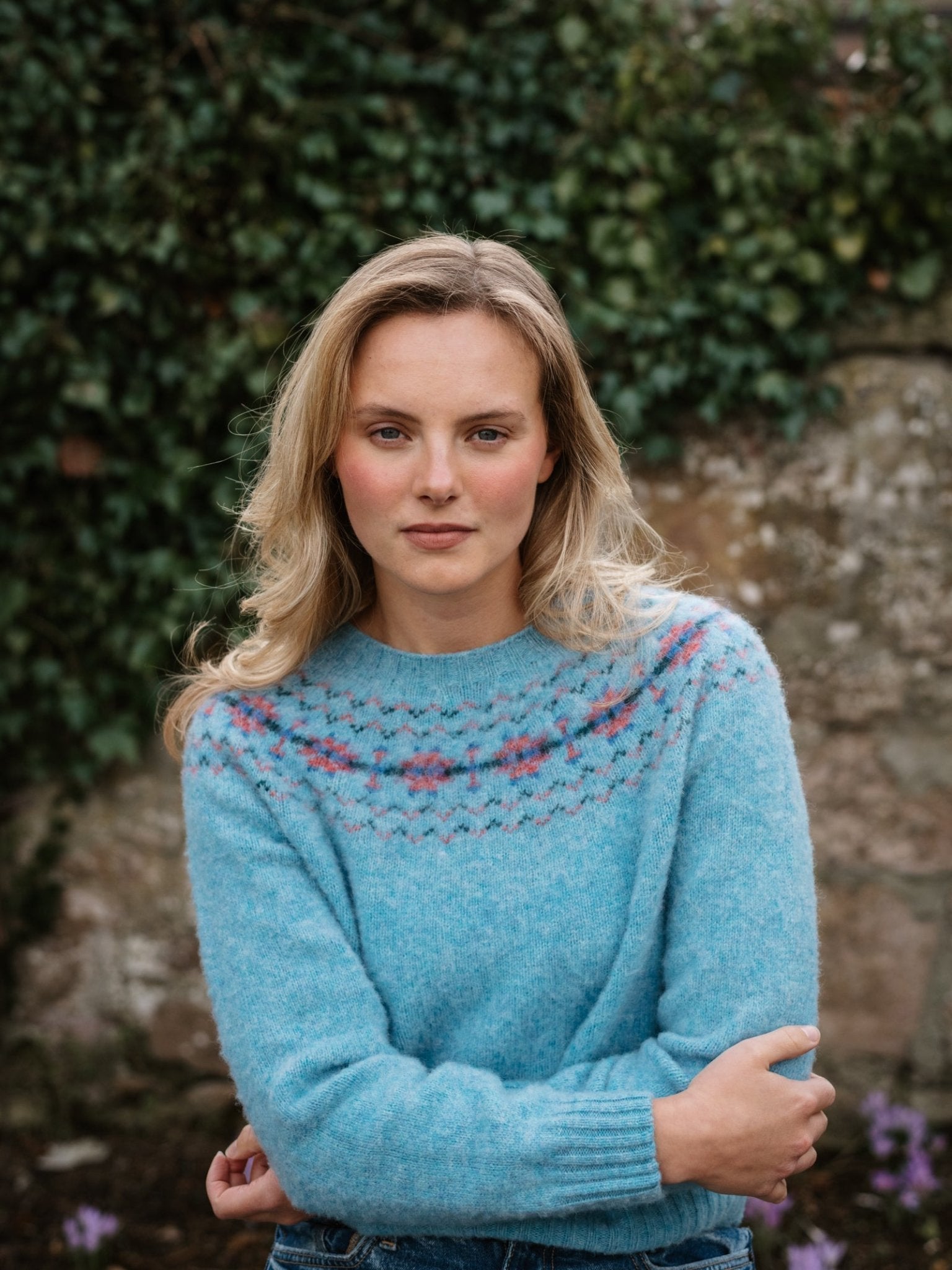 A blond woman wears the Campbell's of Beauly Shaggy Cropped Fairisle Crew Jumper with a pink and white patterned yoke, standing outdoors by an ivy-clad stone wall, looking at the camera with a neutral expression.