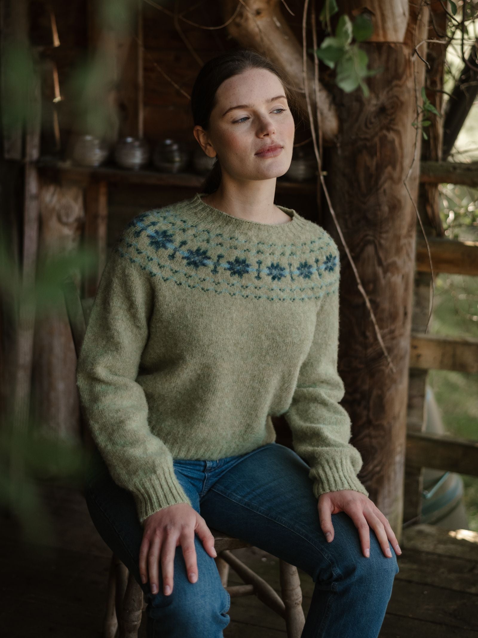 A woman with brown hair in a ponytail sits on a wooden chair, wearing the Campbell's of Beauly Shaggy Cropped Fairisle Crew Jumper in light green and blue, paired with blue jeans, in a rustic sunlit wooden room.