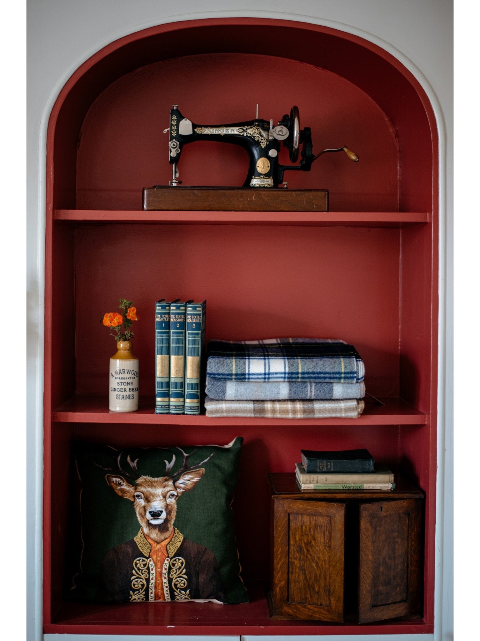 A red arched shelf displays a vintage sewing machine, a vase with flowers, books, folded blankets, and a Campbell's of Beauly Alpine Deer Cushion Cover for a whimsical touch, next to a small wooden cabinet.