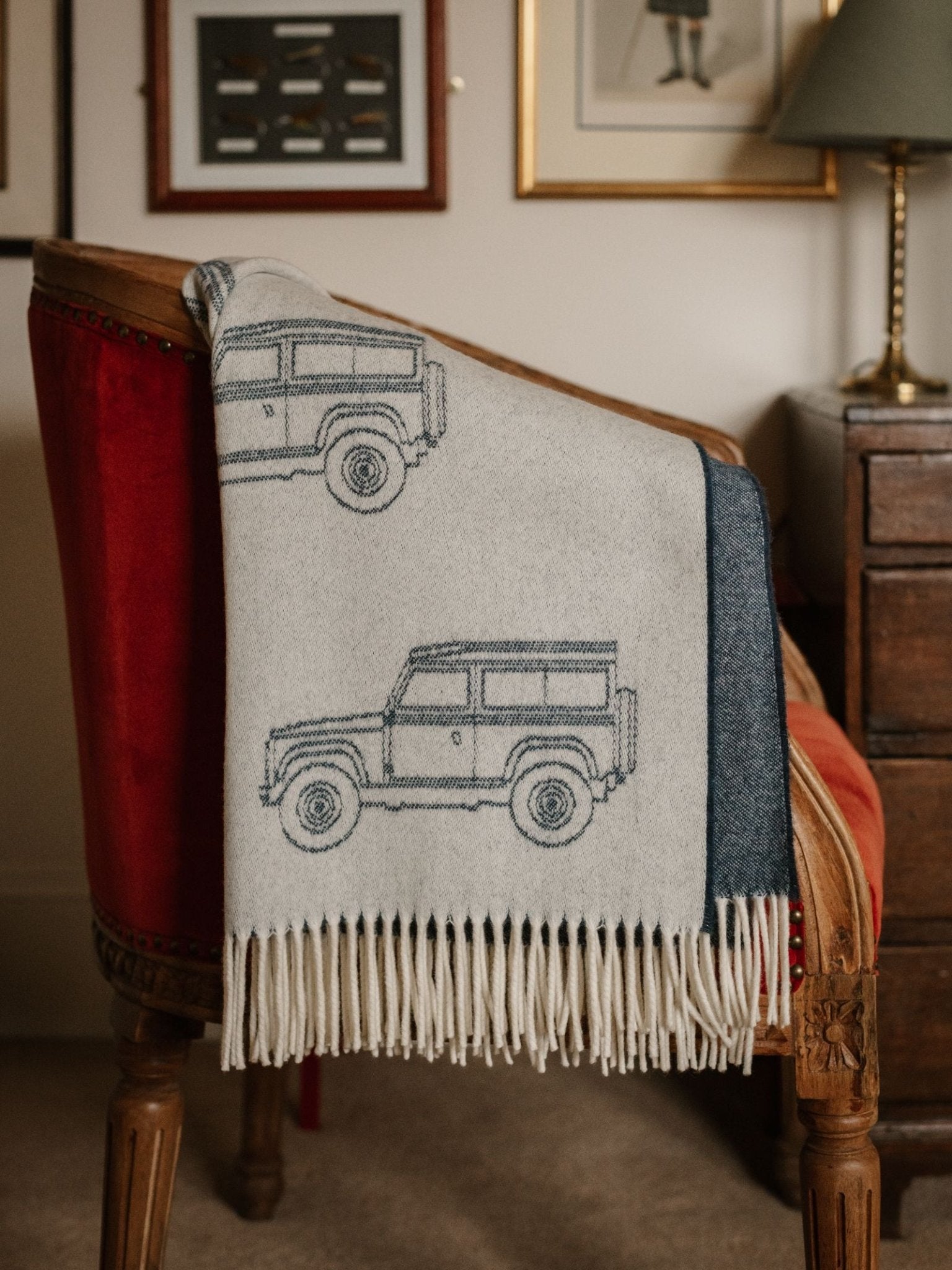 A Land Rover Defender Jacquard Throw by Campbell's of Beauly, featuring outlined illustrations of two classic off-road vehicles, is draped over a wooden armchair with red upholstery beside a wooden desk and wall art.