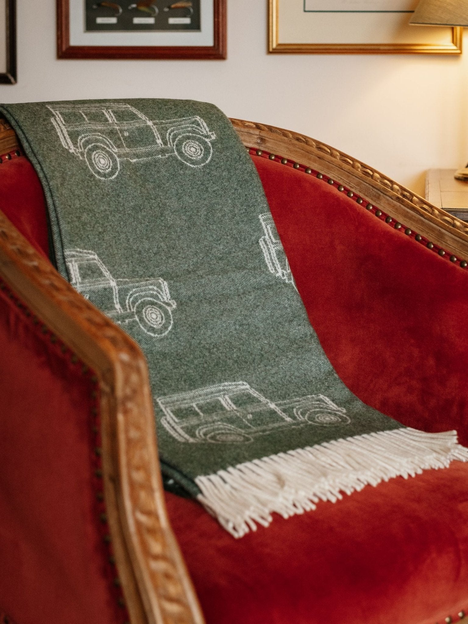 The Land Rover Defender Jacquard Throw by Campbell's of Beauly, in green wool with white car outlines and fringe, is draped over a red velvet armchair with wooden trim in a warmly lit room decorated with framed pictures.