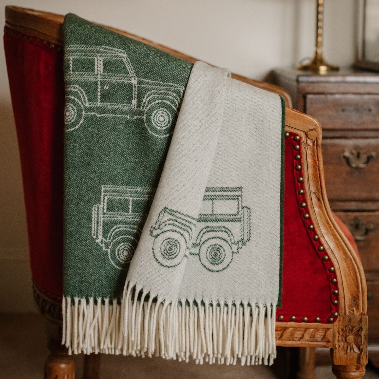 The Campbell's of Beauly Land Rover Defender Jacquard Throw, featuring green and white SUV line drawings, is draped over a vintage red velvet chair beside a wooden dresser in a cozy room.