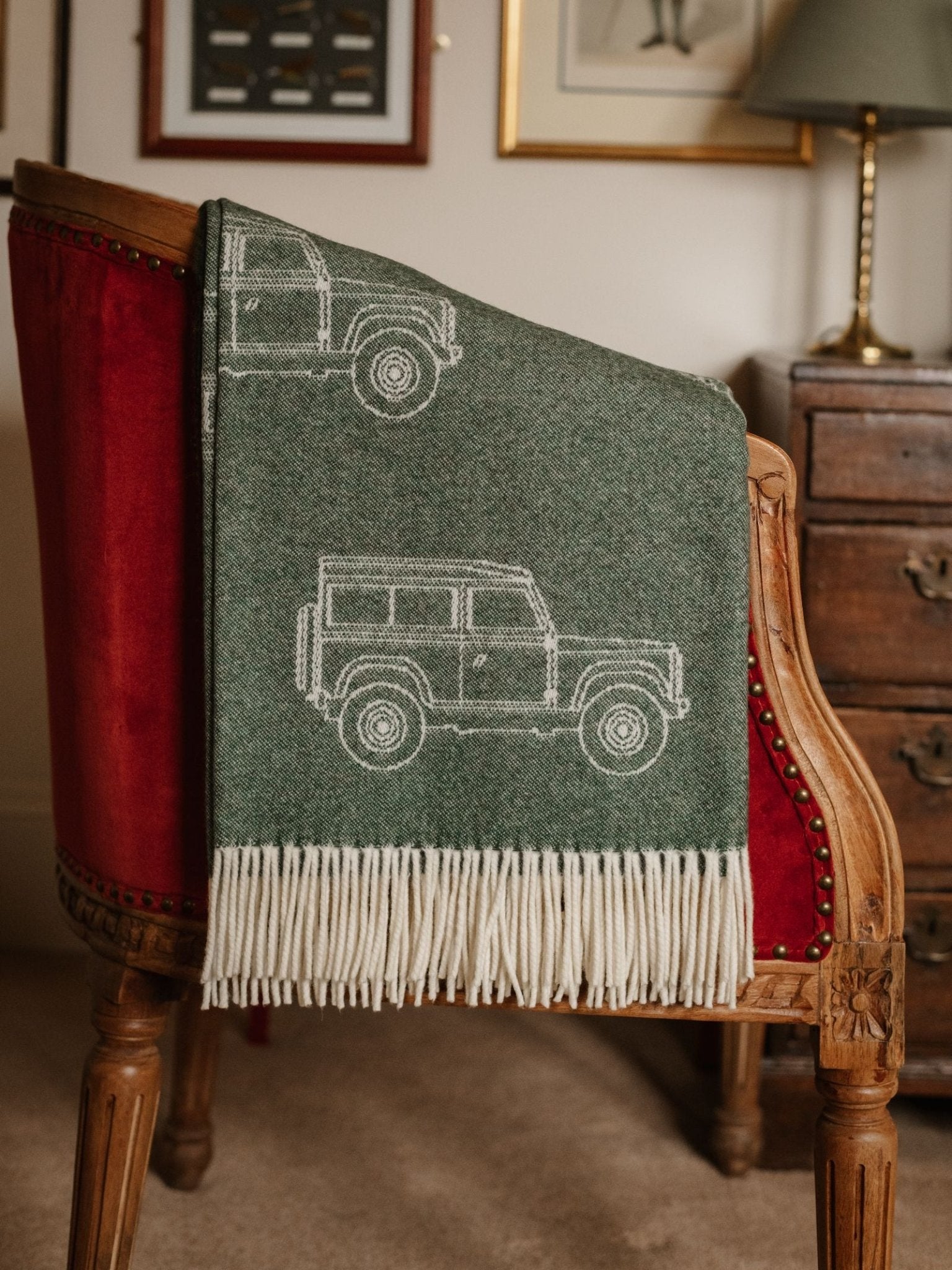 The Land Rover Defender Jacquard Throw by Campbell's of Beauly, a green and white British wool blanket with a classic SUV design, is draped over a vintage wooden chair with red upholstery in a warmly decorated room.