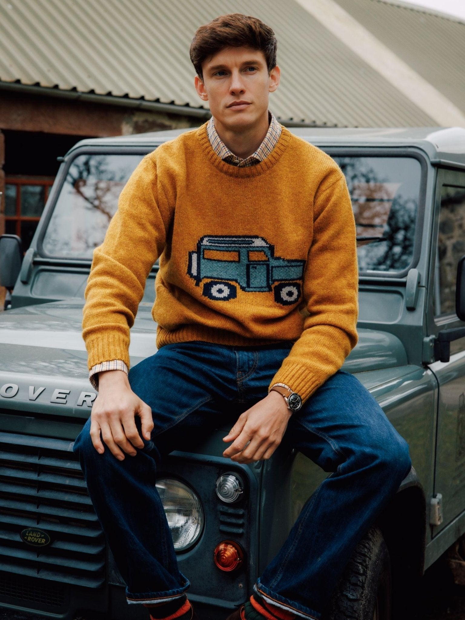 A young man sits on a green Land Rover, wearing Campbell's of Beauly’s Unisex Land Rover Defender Jumper in butterscotch, blue jeans, and a plaid shirt, posing outdoors by a building with a corrugated metal roof.