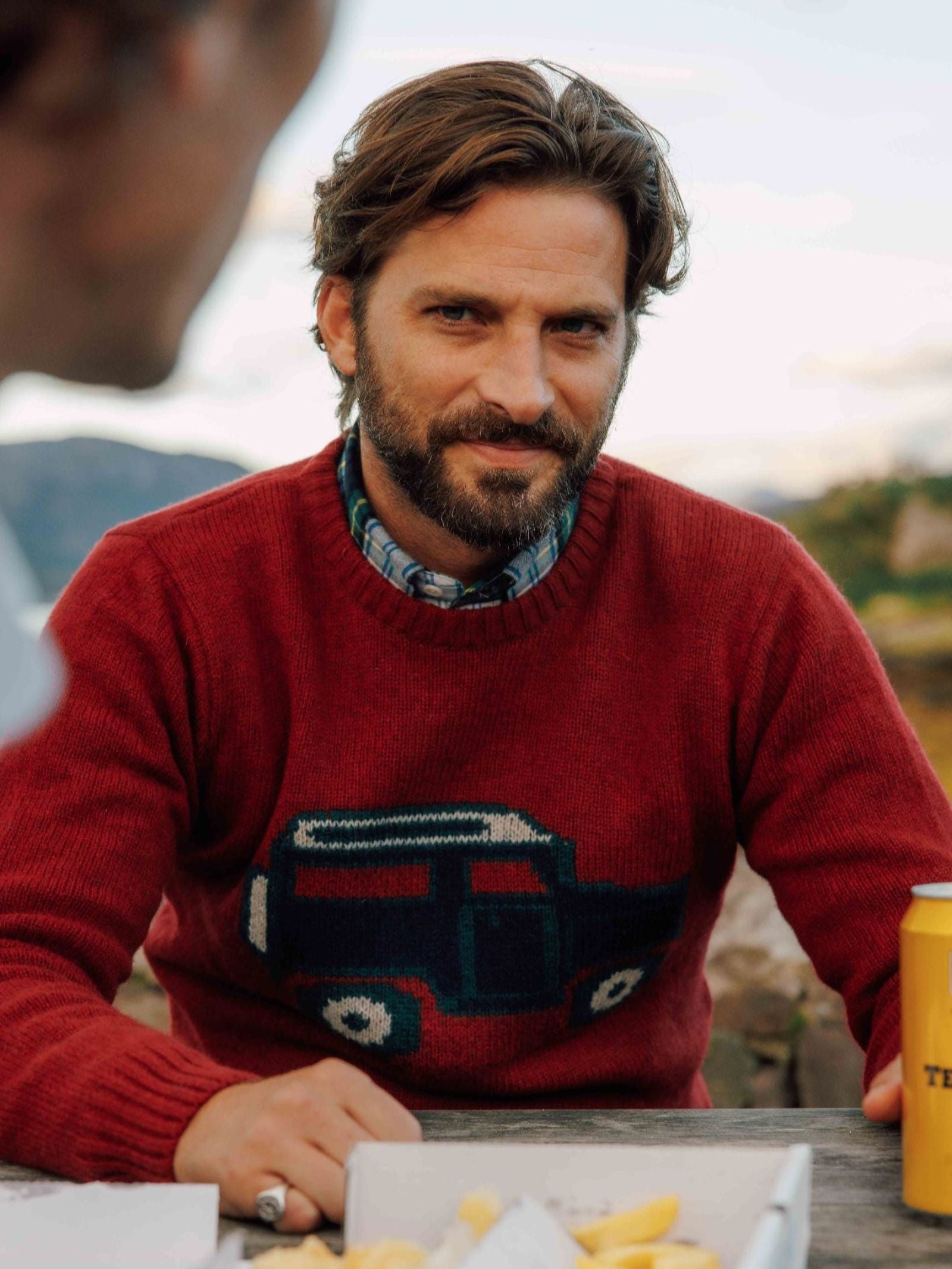 A bearded man in a classic Unisex Land Rover Defender Jumper by Campbell's of Beauly sits outdoors, smiling at the camera with food and a yellow cup on the table; another person is partially visible in the foreground.