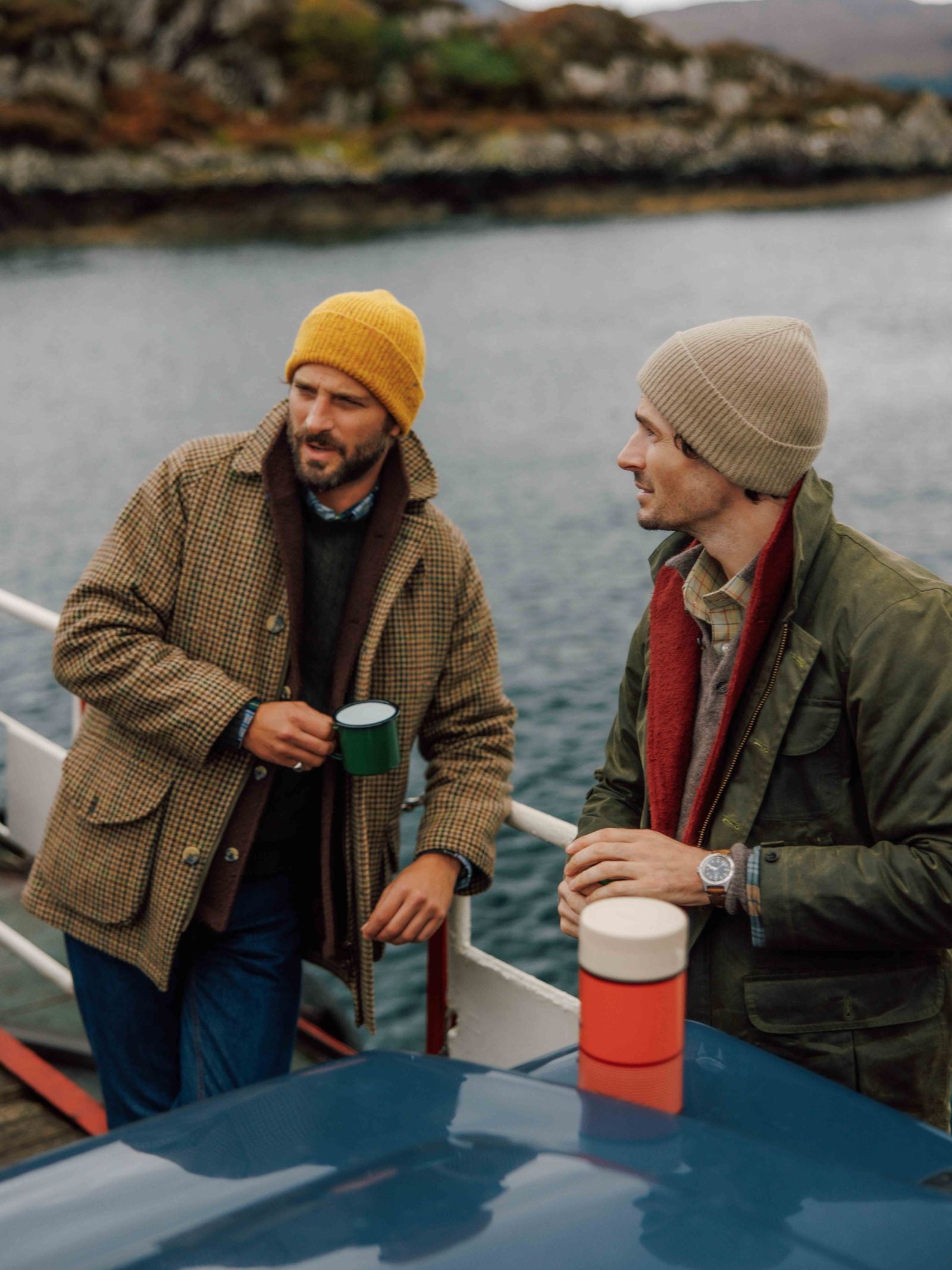 Two men stand by the water holding mugs, wearing Campbell's of Beauly Donegal Lambswool Hats—one in yellow with a brown plaid coat, the other in beige with a green jacket. They look relaxed, chatting outdoors on a dock or boat.