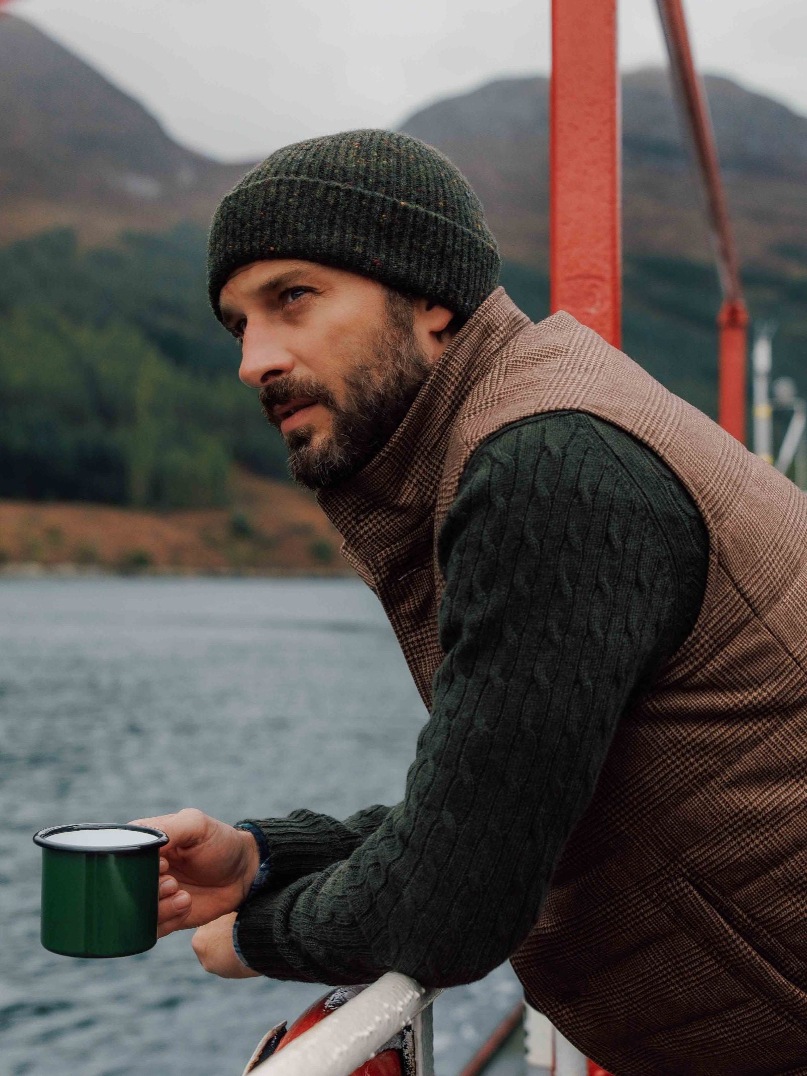 A bearded man in a Campbell’s of Beauly Donegal Lambswool Hat leans on a railing with a green mug, serene lake and forested Scottish Borders mountains in the background.
