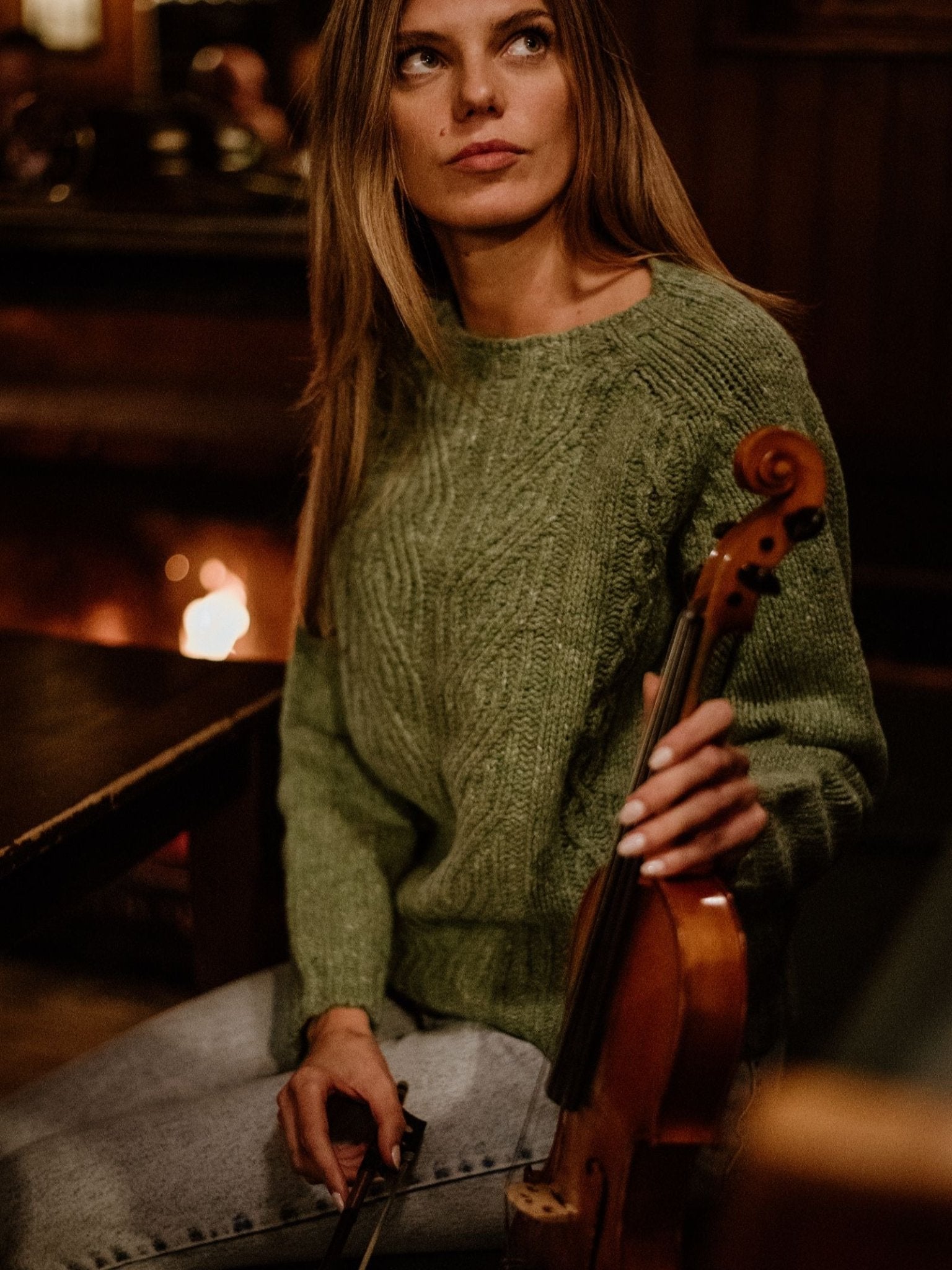 A woman with long hair wears a Campbell's of Beauly Auld Stock Donegal Lambswool Cable Jumper indoors, holding a violin and bow. A warm fireplace glows in the background, creating a cozy atmosphere.