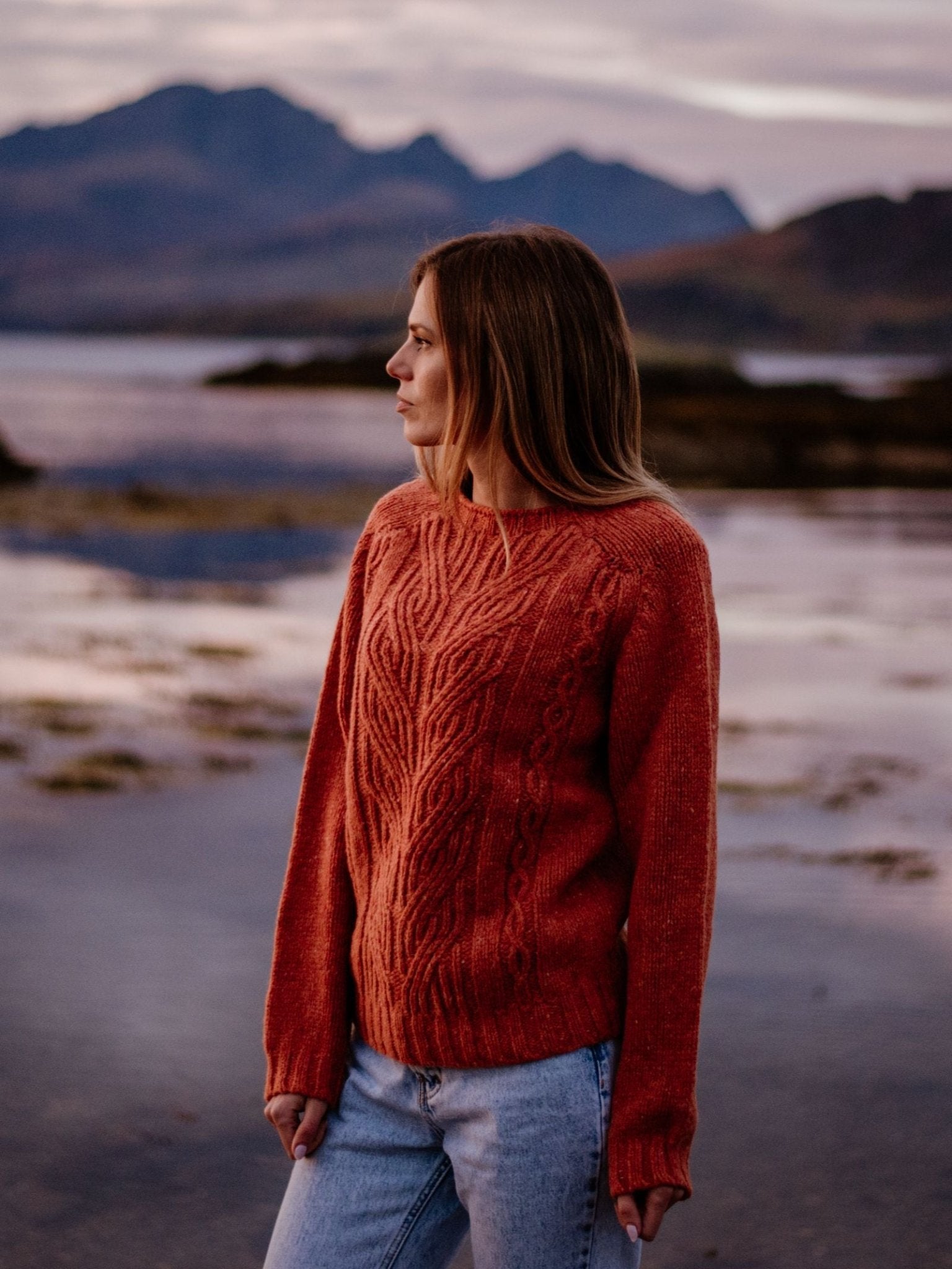 A woman in a burnt orange Campbell's of Beauly Auld Stock Donegal Lambswool Cable Jumper and light blue jeans stands on a beach at sunset, gazing aside with mountains and water behind her.