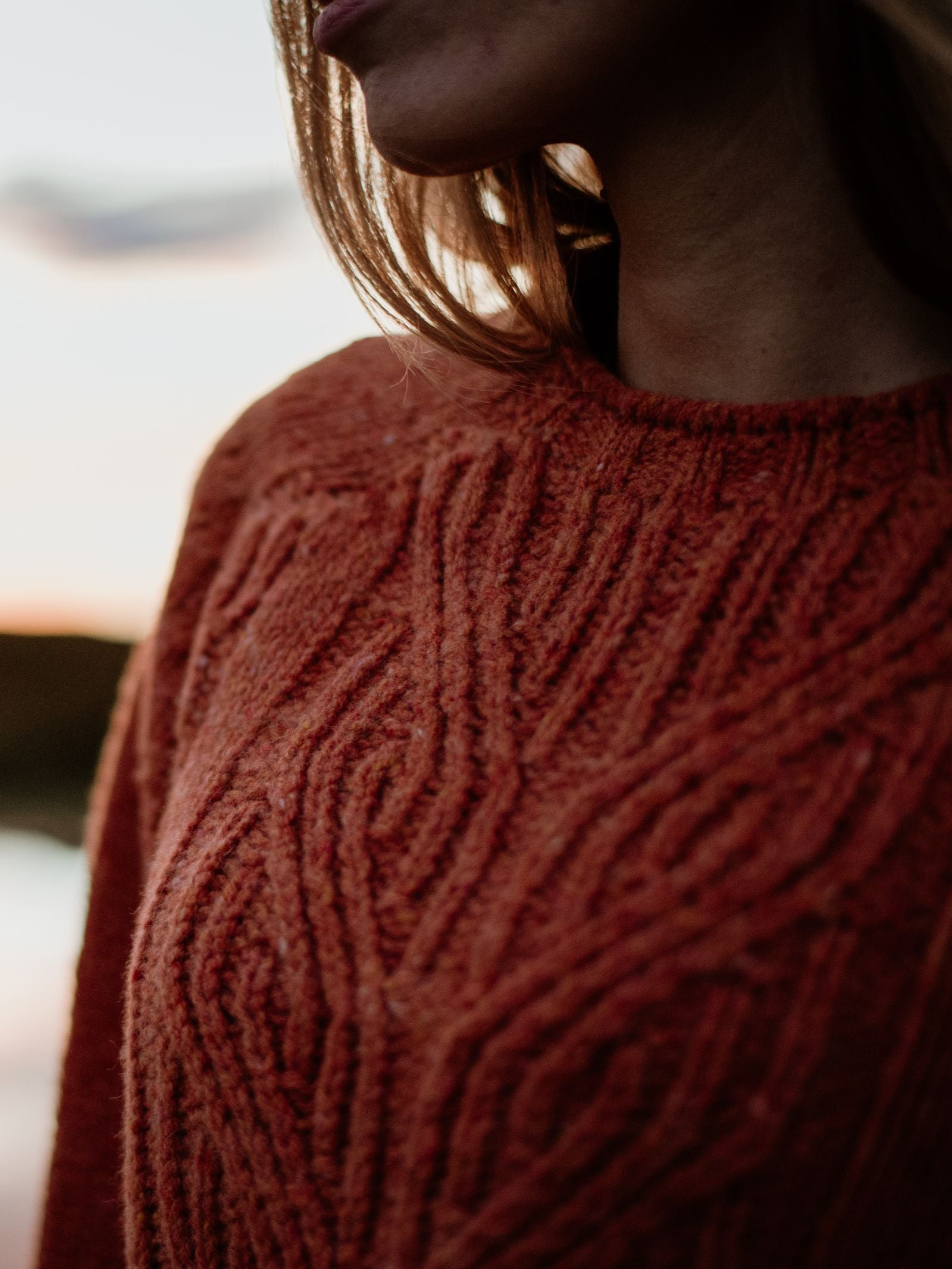 Close-up of a person wearing the Auld Stock Donegal Lambswool Cable Jumper by Campbell's of Beauly in rust, with their face partially out of frame and soft natural light in the background.