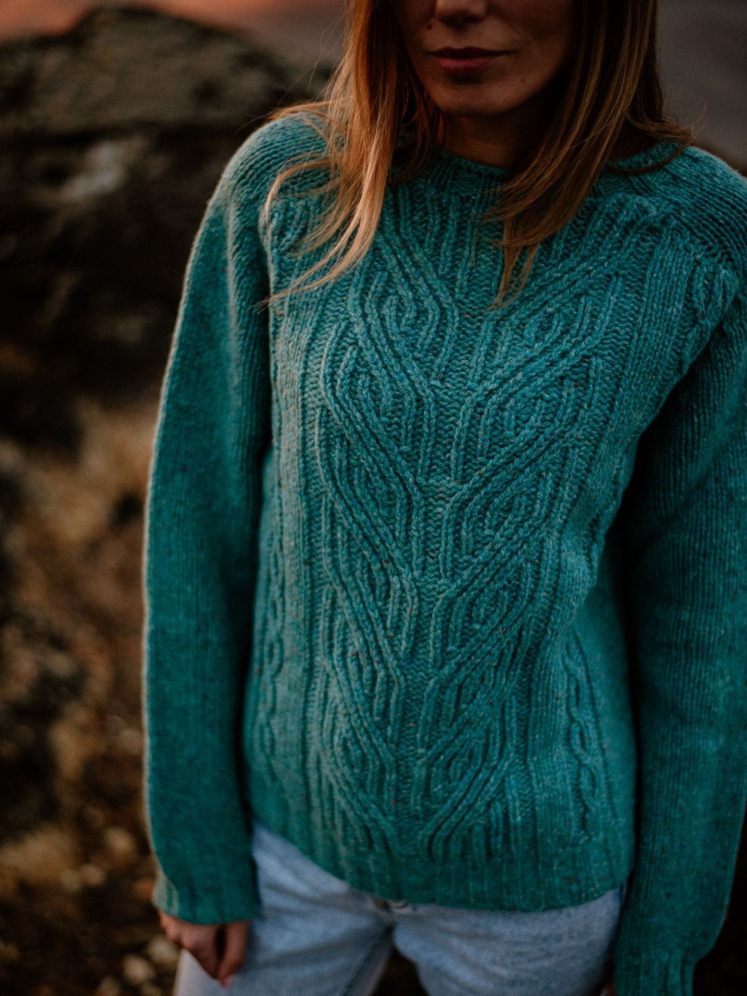A person with long hair wears a teal Campbell's of Beauly Auld Stock Donegal Lambswool Cable Jumper and light pants outdoors by rocks. The shot highlights the jumper’s intricate texture, with their face partially out of frame and in shadow.