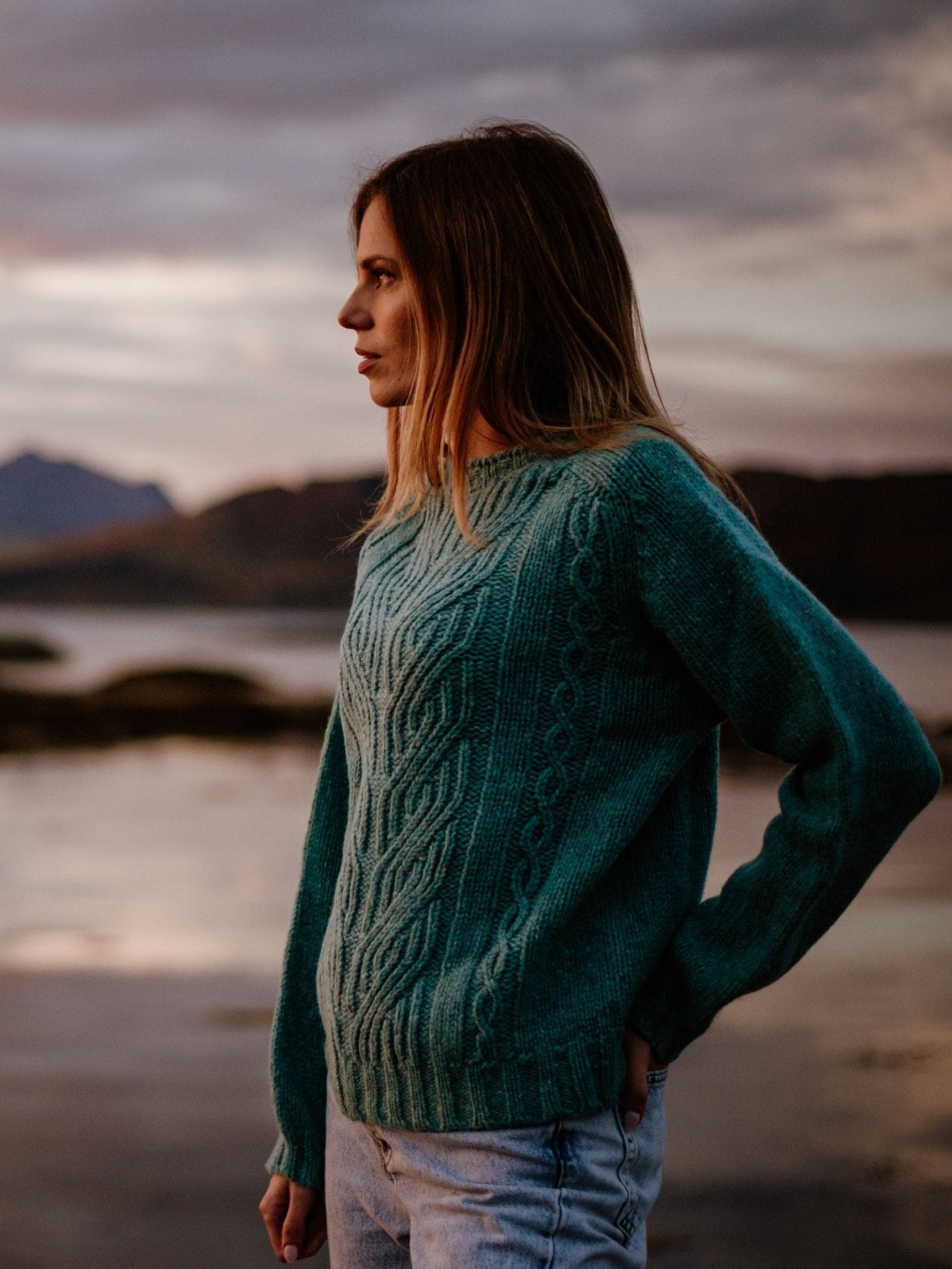 A woman with long blonde hair stands by the water at sunset, wearing the Campbell's of Beauly Auld Stock Donegal Lambswool Cable Jumper in teal and light blue jeans, gazing thoughtfully into the distance.