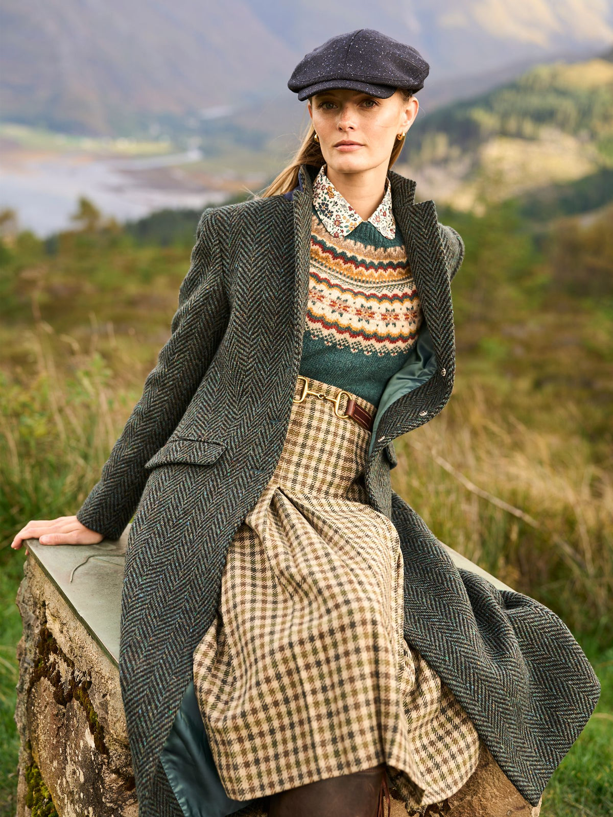 A person wearing a Campbell’s of Beauly Dogtooth Skirt, a fair isle knit sweater, and a herringbone overcoat, sitting on a stone wall in a rural landscape.