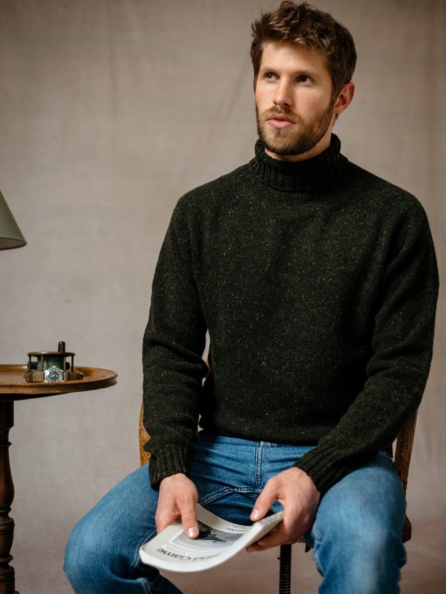 A man with short brown hair and a beard, wearing Campbell's of Beauly Donegal Lambswool Roll Neck sweater and blue jeans, sits on a chair holding a folded newspaper. A small round table with a lamp and clock is beside him.