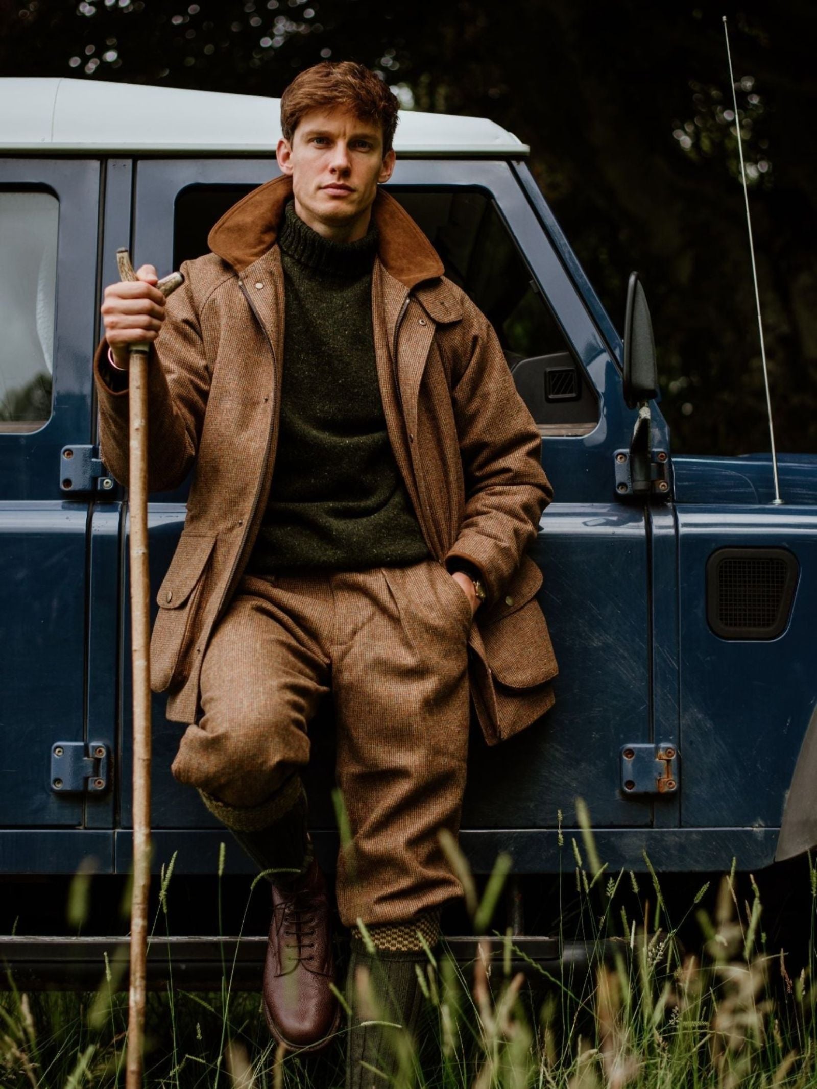 A man wearing a Campbell's of Beauly Donegal Lambswool Roll Neck and brown tweed leans on a blue off-road vehicle, holding a wooden walking stick, with tall grass and trees in the background.