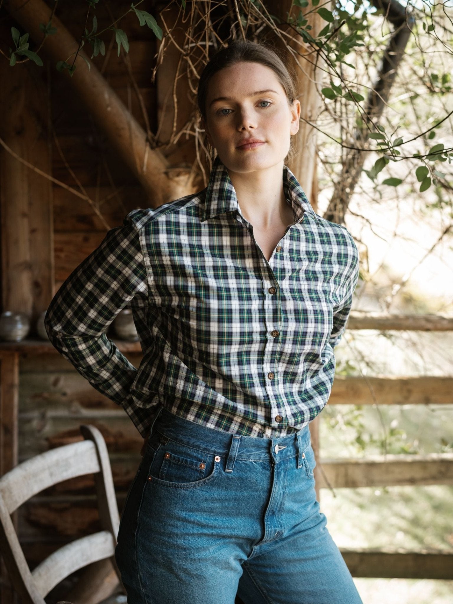A woman stands on a wooden porch in blue jeans and the Cotton Tartan Shirt by Campbell's of Beauly, hands behind her back. She faces the camera, framed by rustic wooden walls and leafy branches.