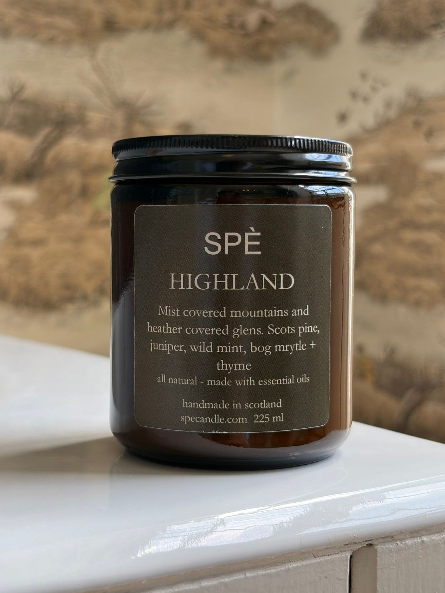 A brown glass candle jar labeled SPÈ Atelier Highland Candle sits on a white surface with a patterned background. The label lists natural ingredients and notes that it is handmade in Scotland, featuring a botanical scent inspired by the juniper-laced air of the Scottish Highlands.