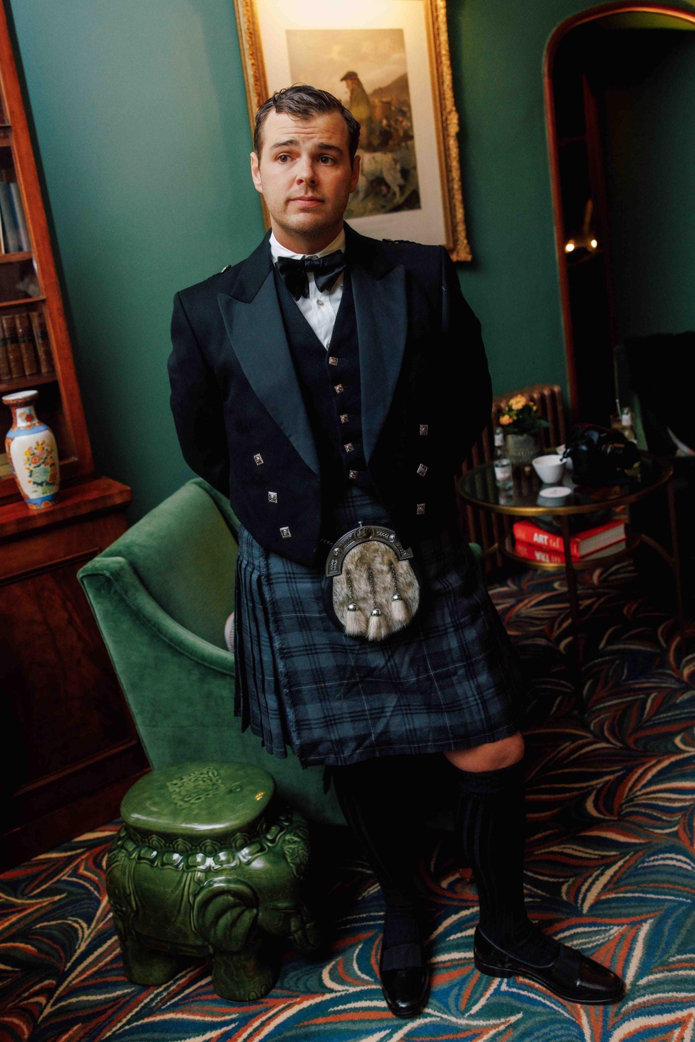 A man dressed in traditional Scottish attire, including a black jacket, bow tie, kilt, and sporran, stands in a stylish room with green walls, a green armchair, and patterned carpet.