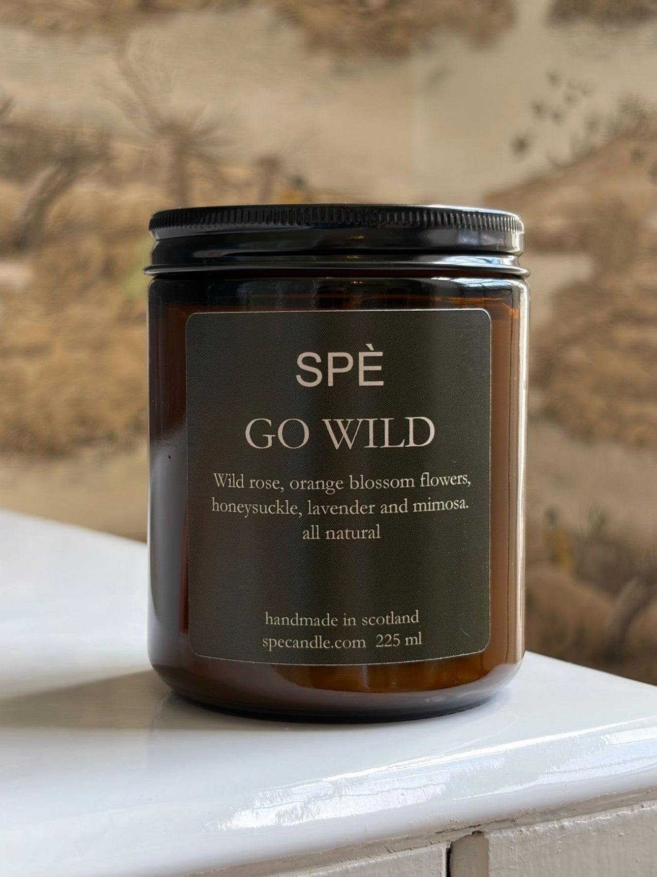 A brown glass candle jar labeled "Go Wild Candle" by SPÈ Atelier rests on a white surface, set against beige nature-themed wallpaper, offering soft floral notes and green botanicals for a refreshing ambiance.