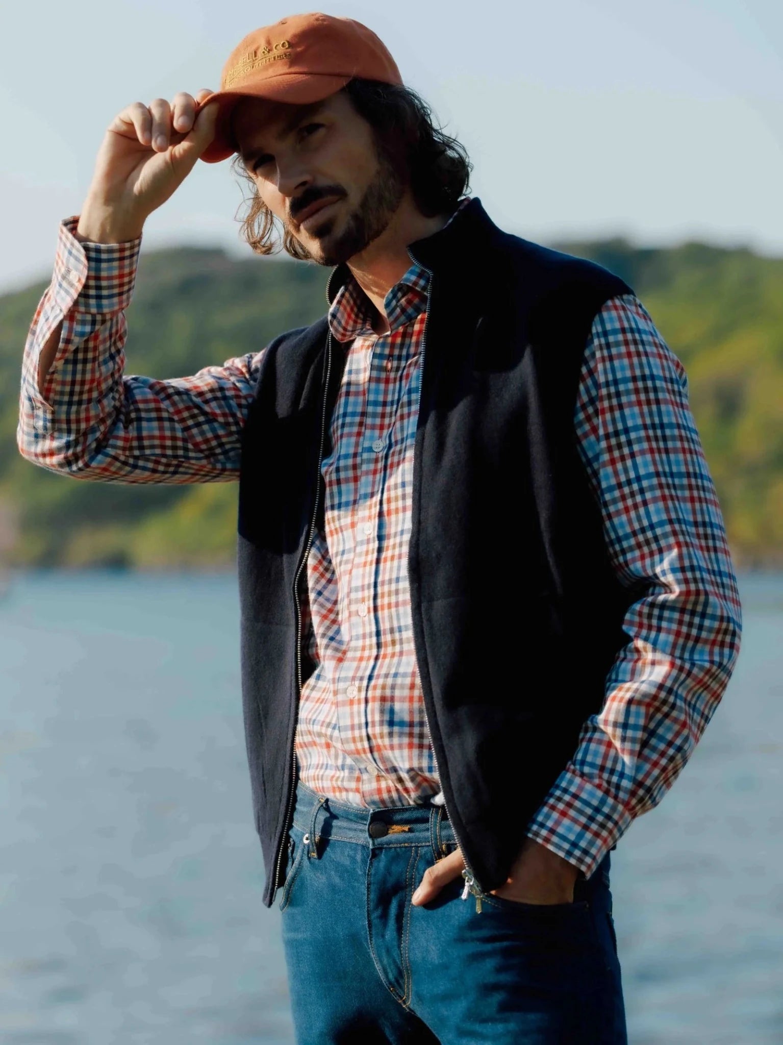 A man in an orange cap, navy vest, jeans, and the Campbell’s of Beauly Auld Stock Windowpane Shirt stands outdoors by water, one hand on his cap and one in his pocket, with trees and a hillside behind him.