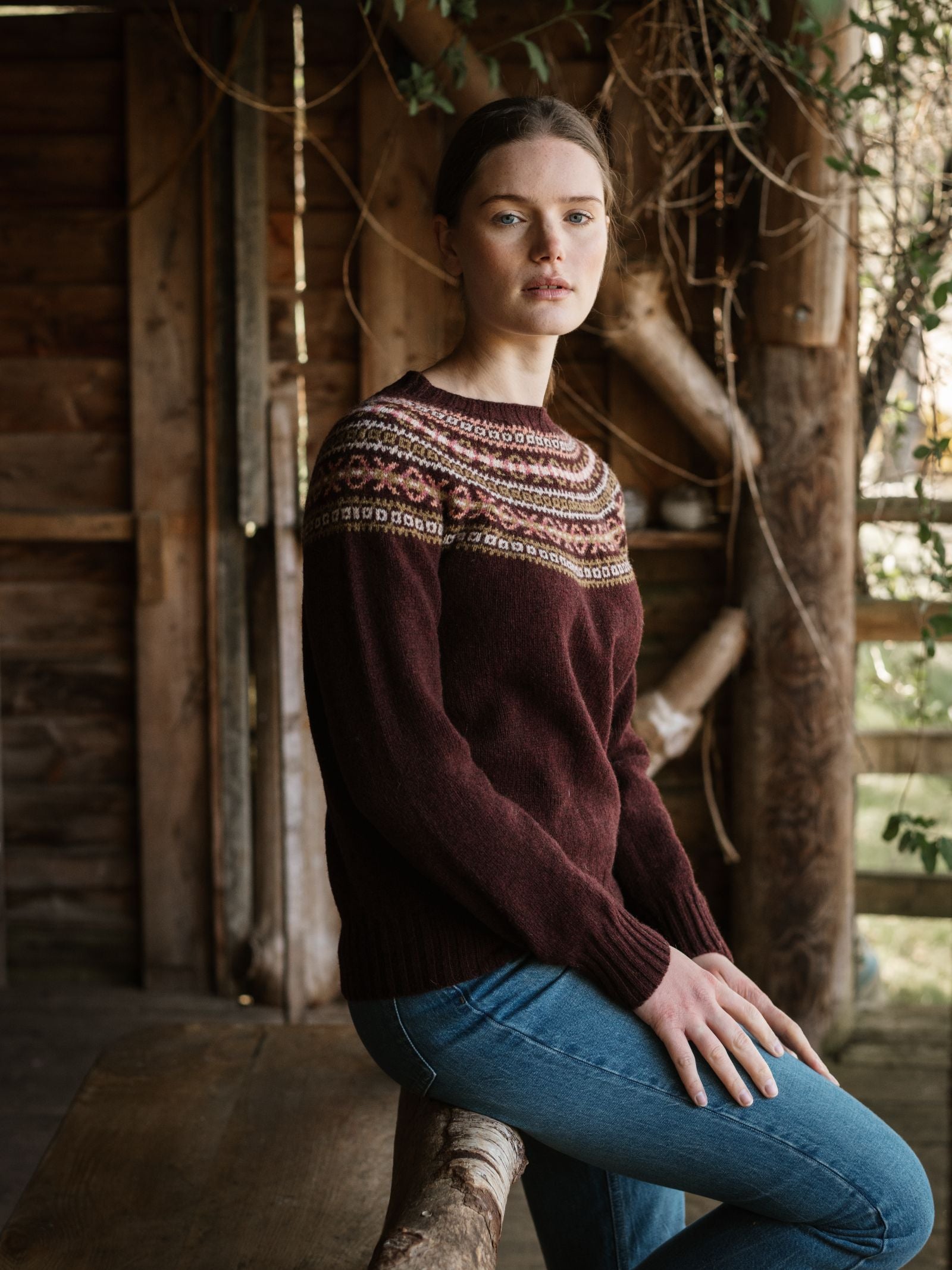 A woman with light skin and brown hair, wearing the Campbell's of Beauly Fairisle Crew Jumper and blue jeans, sits calmly on a wooden bench indoors. Rustic wooden walls and greenery form the background.