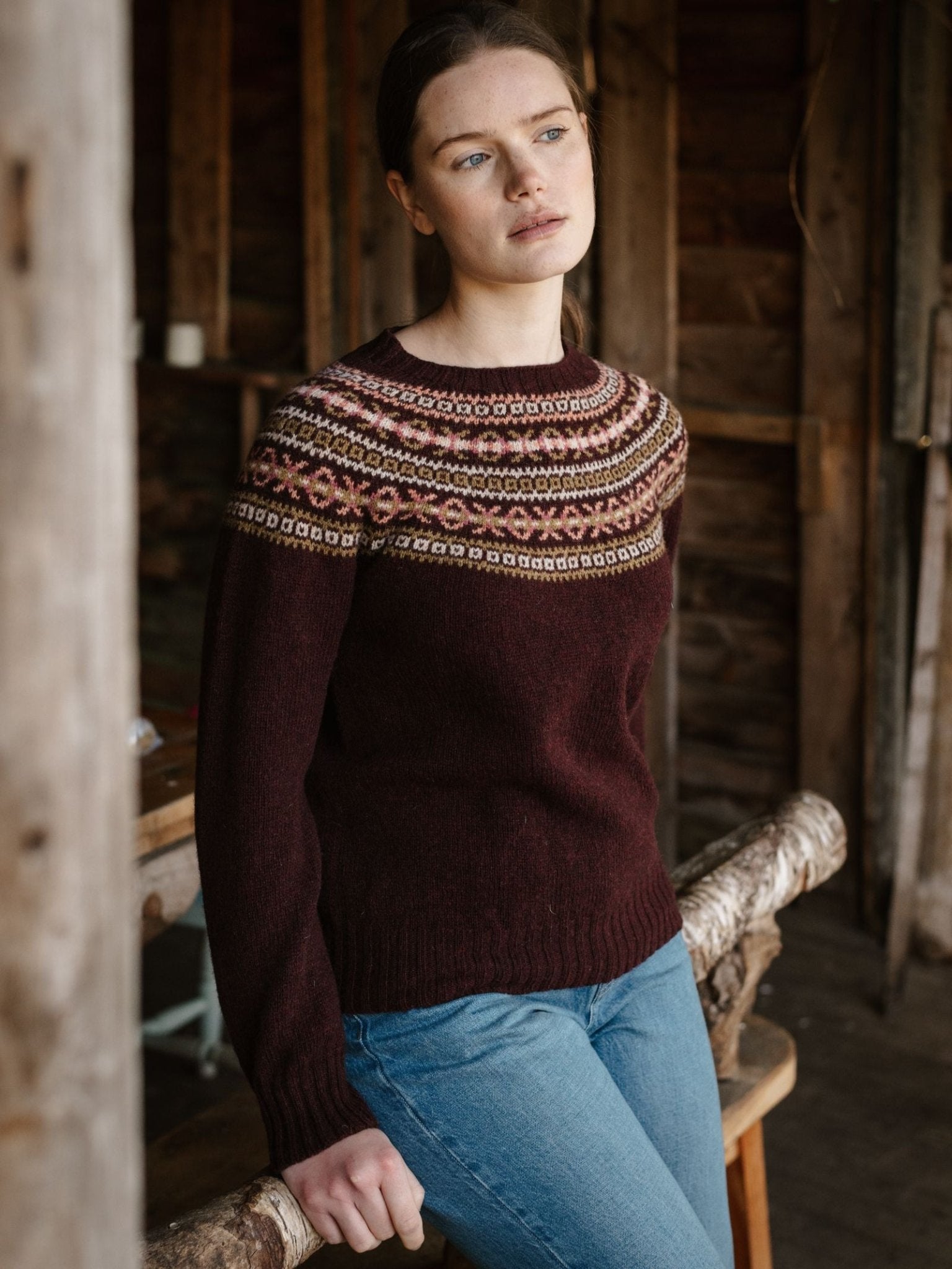 A woman in Campbell's of Beauly Fairisle Crew Jumper, featuring a maroon Geelong lambswool knit with a colorful yoke, leans against a wooden beam inside a rustic building, gazing thoughtfully ahead. She pairs it with blue jeans and has her hair pulled back.