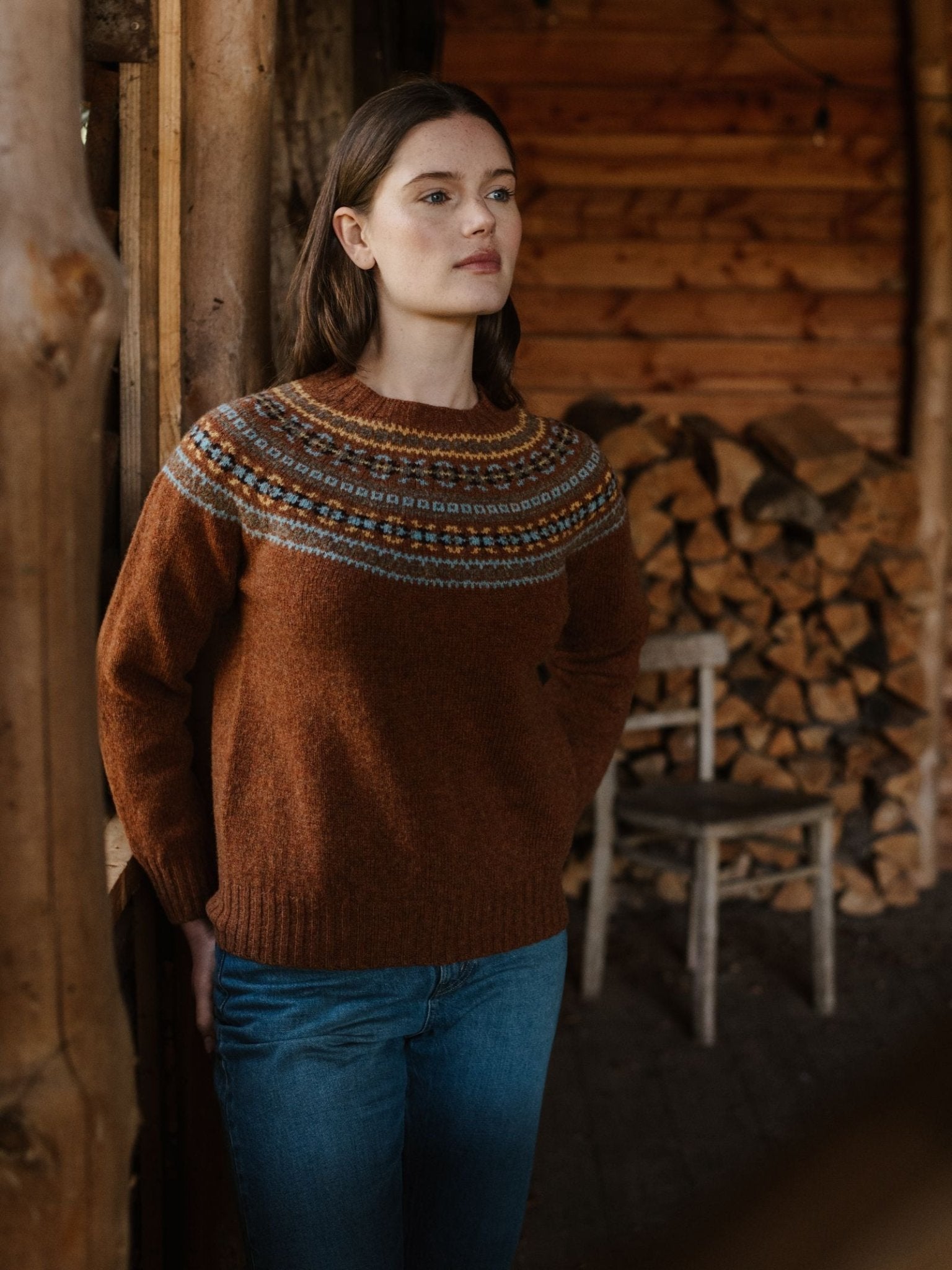 A woman in a brown patterned Fairisle Crew Jumper by Campbell's of Beauly and blue jeans stands indoors near stacked firewood and wooden walls, with a rustic chair in the background.