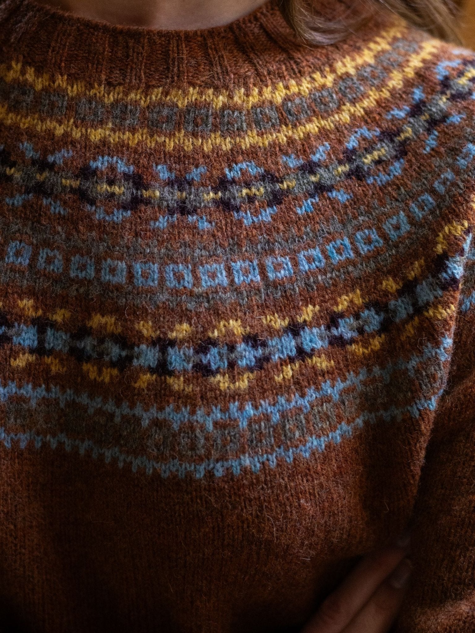 A close-up of a person wearing the Campbell's of Beauly Fairisle Crew Jumper, crafted in Scotland from soft Geelong lambswool, featuring intricate blue, yellow, and white patterns across the chest and shoulders, with arms crossed.