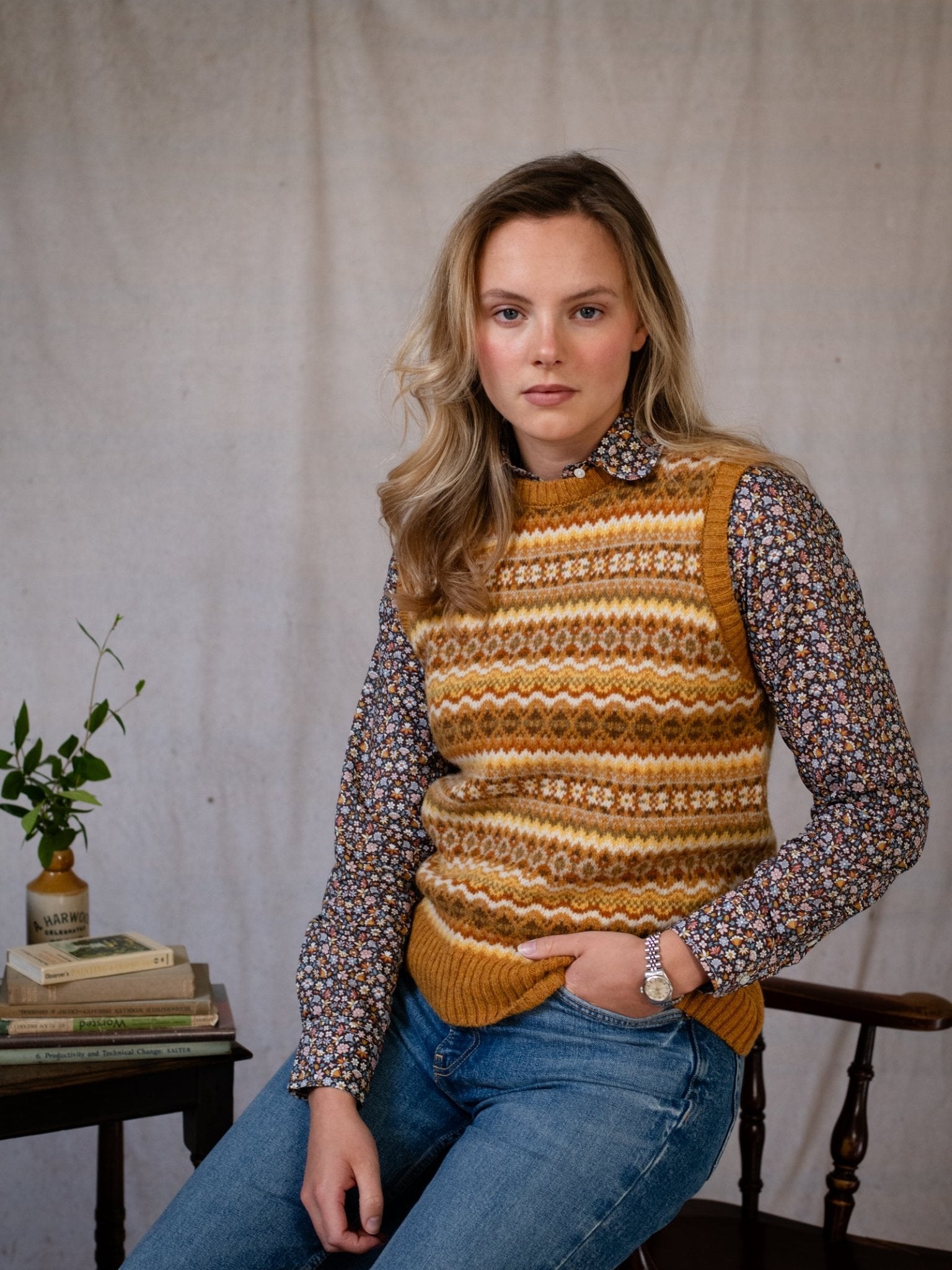 A woman with blonde hair sits on a wooden chair, wearing a Campbell's of Beauly Fairisle Crew Tank, knitted in Scotland from soft Shetland wool, over a floral shirt and blue jeans. She rests her hand on a table with books and greenery against a neutral backdrop.