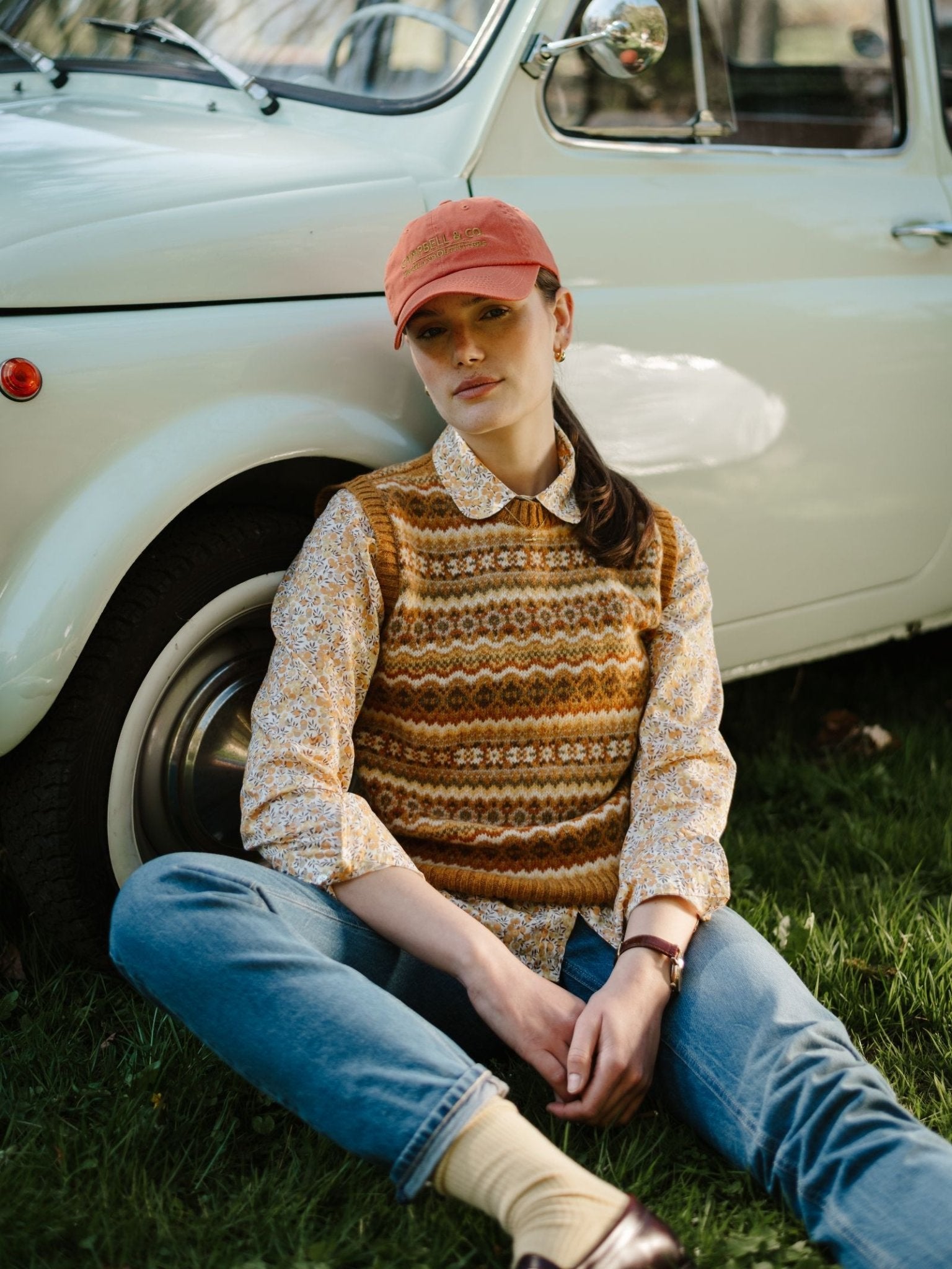 A woman in a patterned sweater vest, Campbell's of Beauly Liberty Print Cotton Shirt, jeans, and a pink cap sits on grass, leaning against the wheel of a vintage white car.