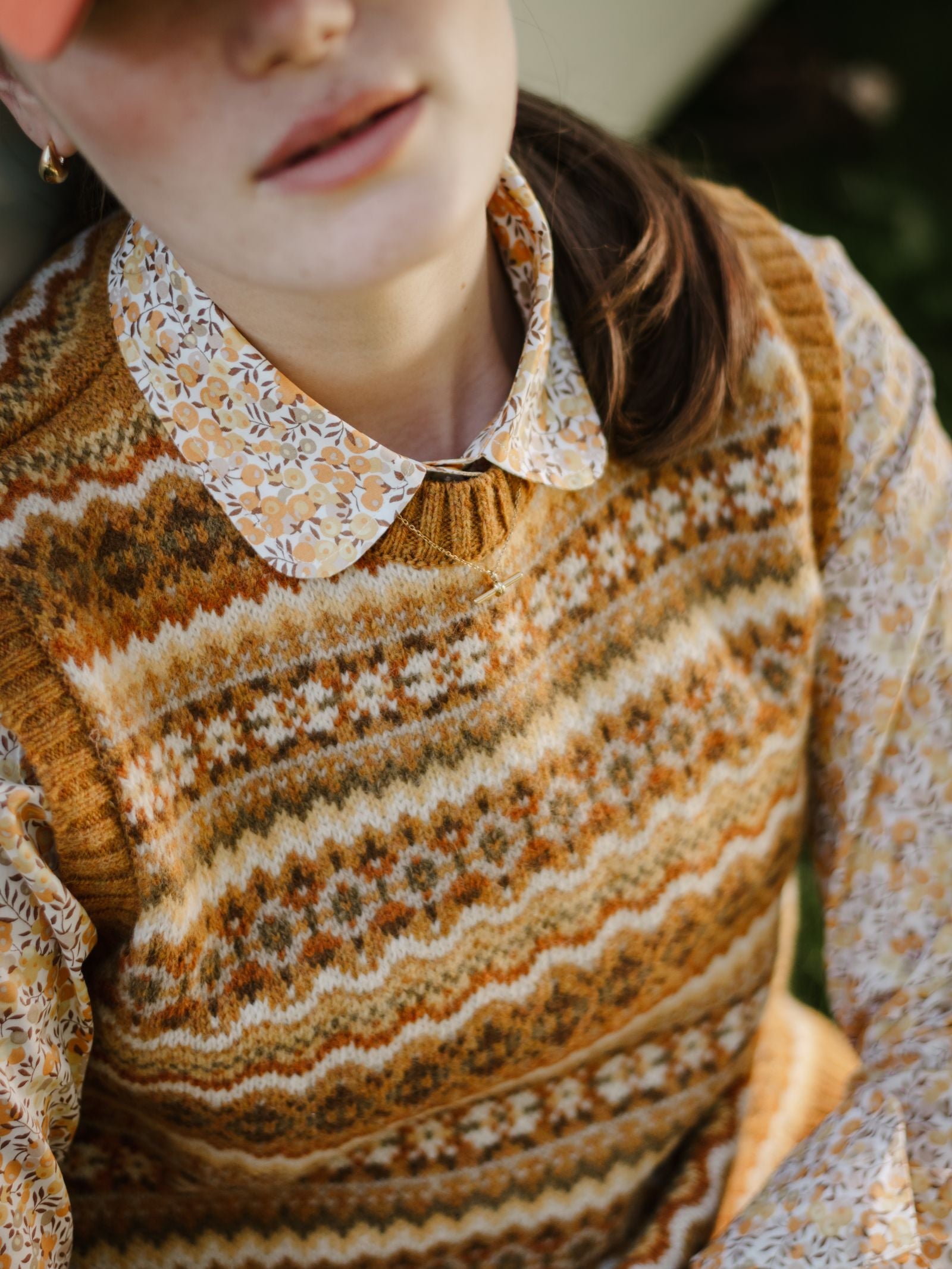A person in a Campbell's of Beauly Liberty Print Cotton Shirt under a patterned brown and yellow knit sweater, with a pink cap and ponytailed brown hair, is sitting outdoors. The image is cropped between the mouth and nose.