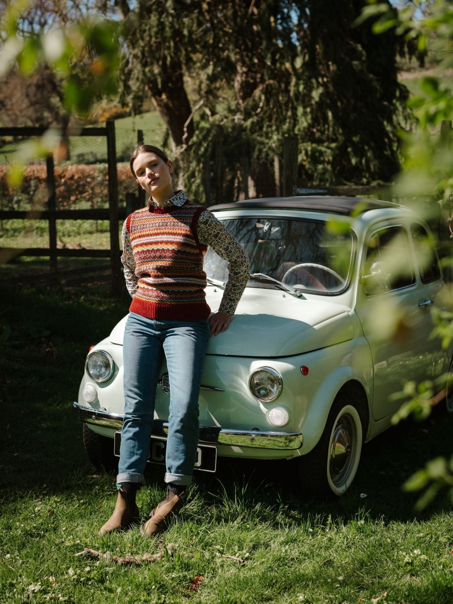 Wearing a Campbell's of Beauly Fairisle Crew Tank over a floral shirt with jeans and boots, someone leans against a vintage white car in a sunlit, grassy yard bordered by trees and a wooden fence.