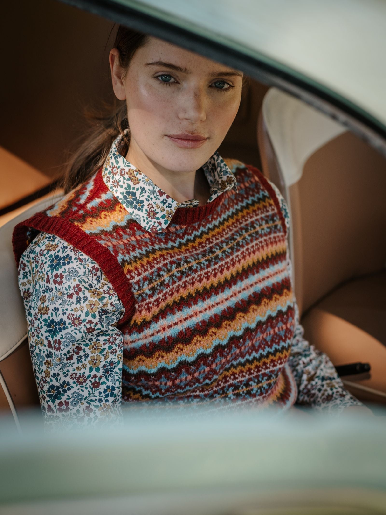A woman sits in a car, looking at the camera. She wears a floral shirt and a Campbell's of Beauly Fairisle Crew Tank, knitted in Scotland from soft Shetland wool. The focus is on her upper body and face.