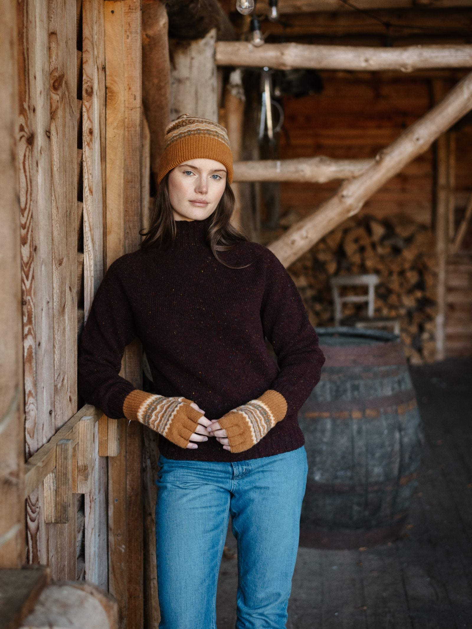 A woman in a knit hat, dark sweater, and Campbell's of Beauly Lambswool Fairisle Half-Finger Gloves in Butterscotch Fairisle leans against a wooden wall inside a rustic cabin with stacked firewood and a barrel behind her.