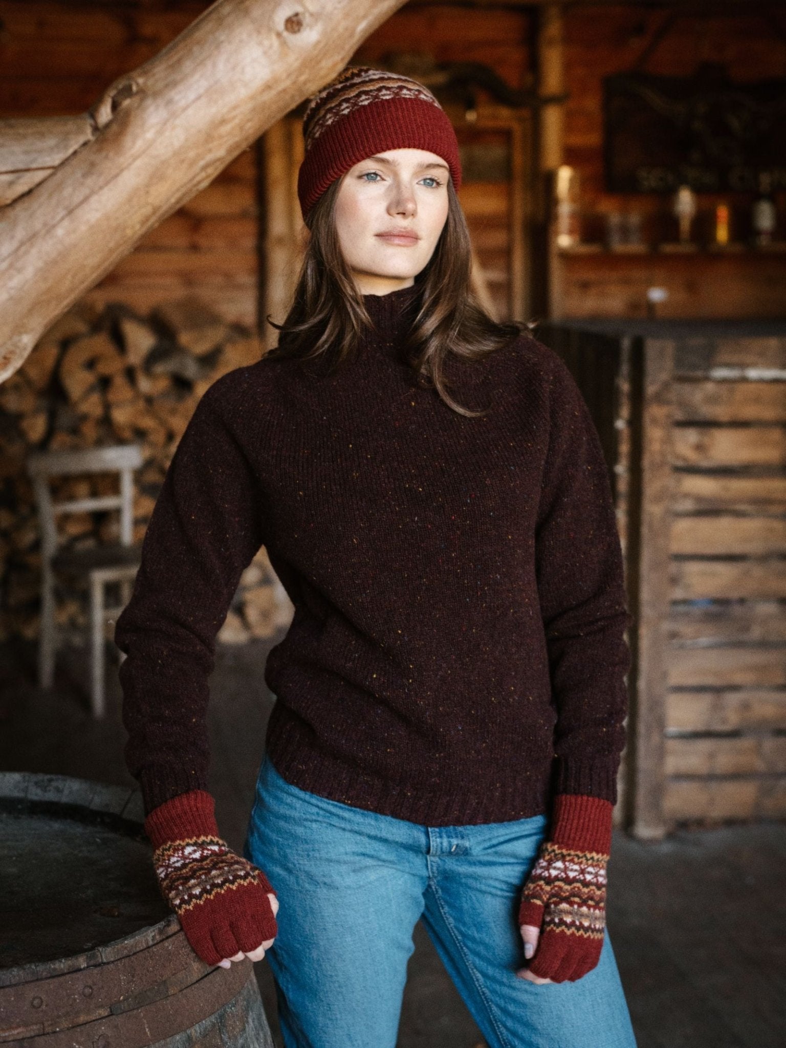 A woman indoors near wooden beams and firewood showcases cozy Scottish knitwear style, wearing blue jeans, a dark brown sweater, and Campbell's of Beauly Lambswool Fairisle Half-Finger Gloves in Sienna Fairisle.