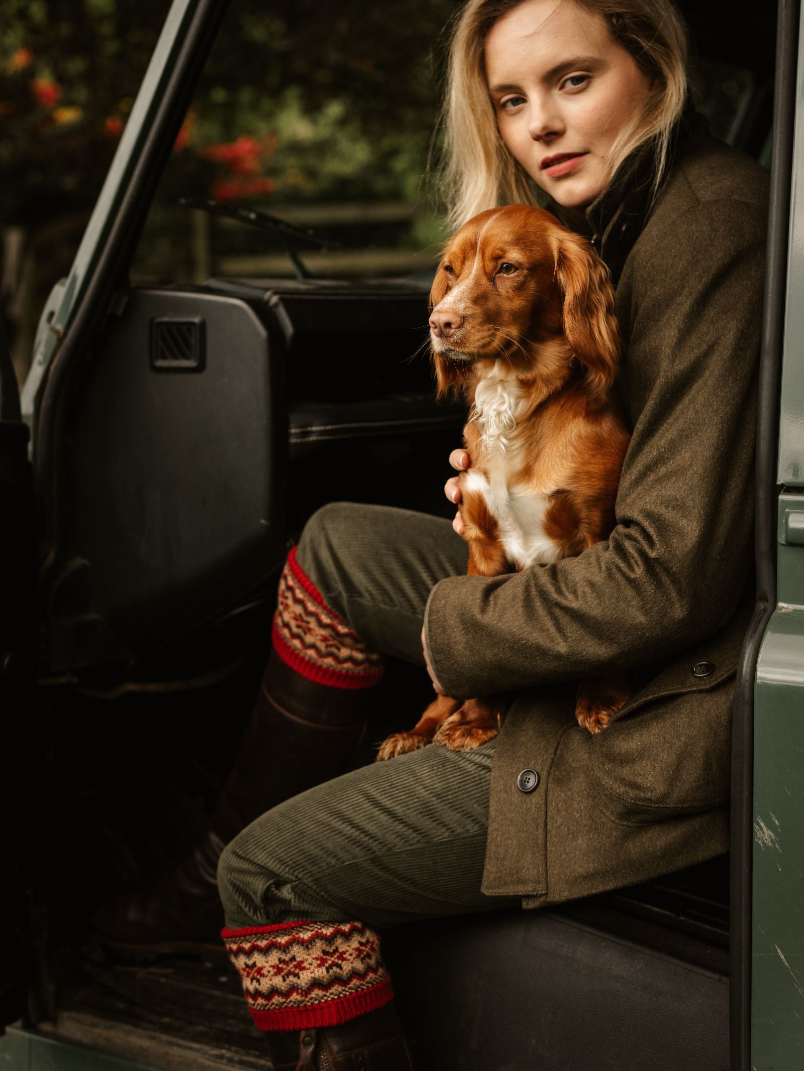 A woman in an olive green jacket and Campbell's of Beauly Merino Fairisle Shooting Socks sits in a car doorway with a small brown dog on her lap, gazing at the camera amid blurred greenery and hints of classic Scottish knitwear.