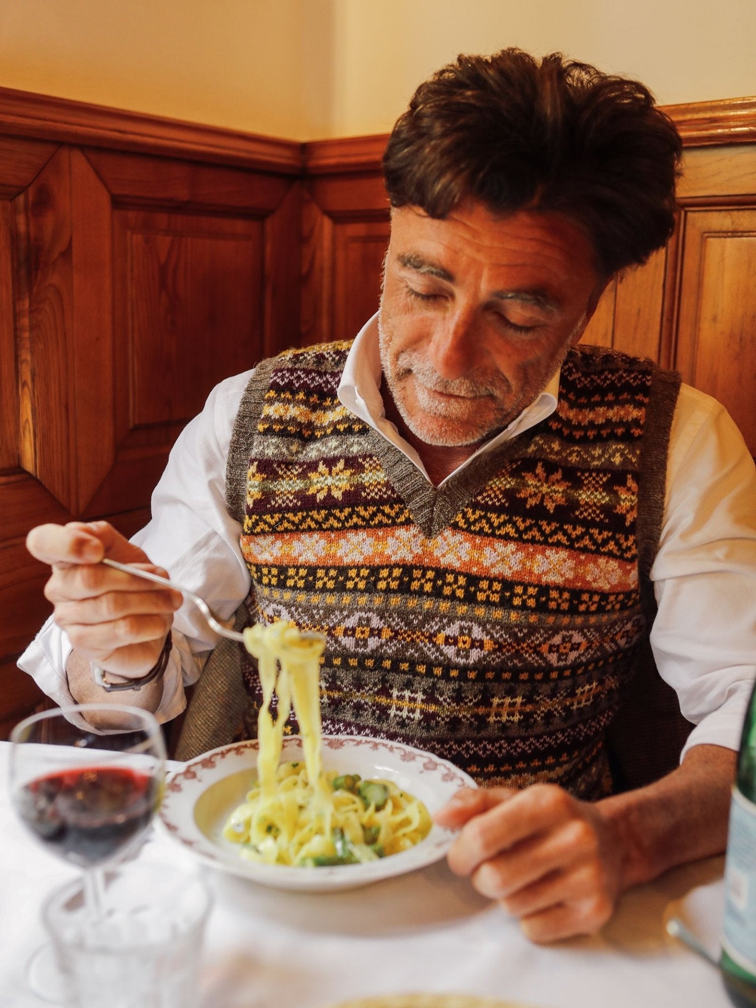 A man wearing the Campbell’s of Beauly Fairisle Slipover—a timeless, sleeveless piece crafted from Shetland wool—sits at a table, twirling pasta onto his fork from a bowl. On the table are a glass of red wine and a bottle of water. The cozy scene is set against walls with wooden paneling.