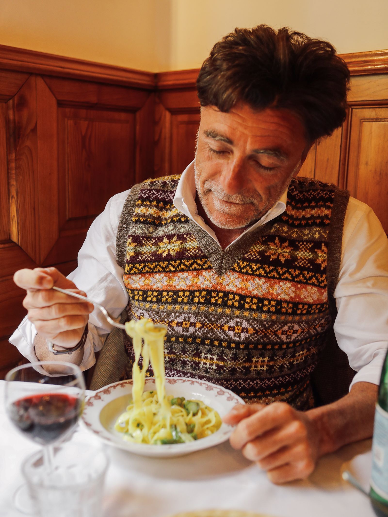 A man wearing the Campbell’s of Beauly Fairisle Slipover—a timeless, sleeveless piece crafted from Shetland wool—sits at a table, twirling pasta onto his fork from a bowl. On the table are a glass of red wine and a bottle of water. The cozy scene is set against walls with wooden paneling.