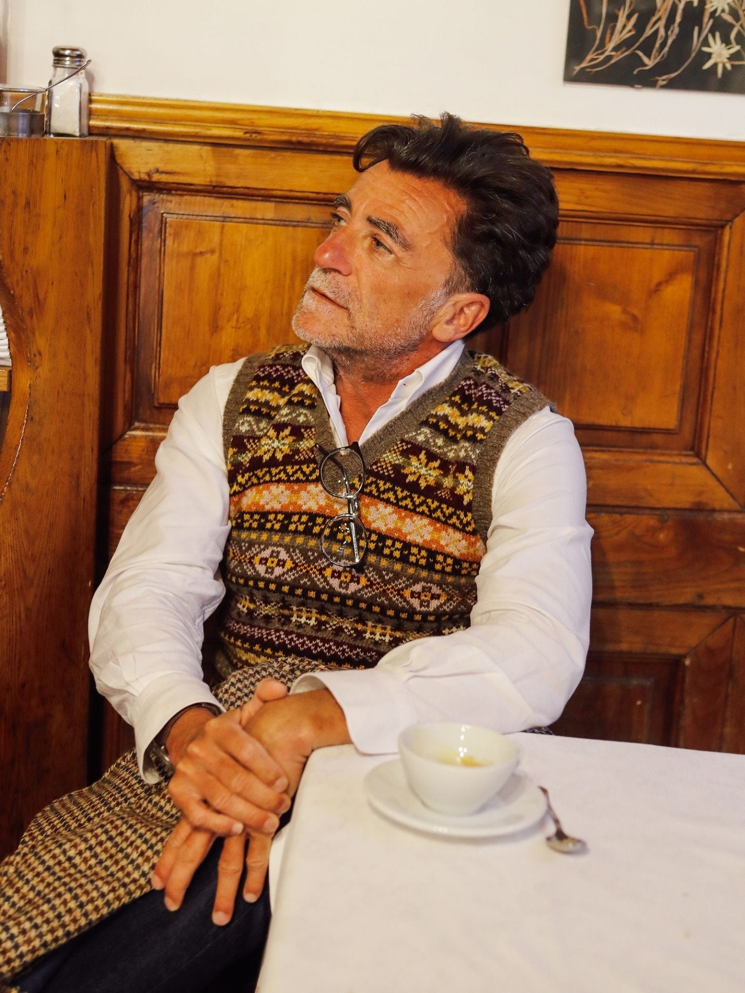 A man with dark hair and a white shirt wears the classic Fairisle Slipover from Campbell’s of Beauly, its Shetland wool lending rich texture to the patterned sweater vest. He sits at a table with a cup and saucer, looking thoughtfully to the side in a wood-paneled room.