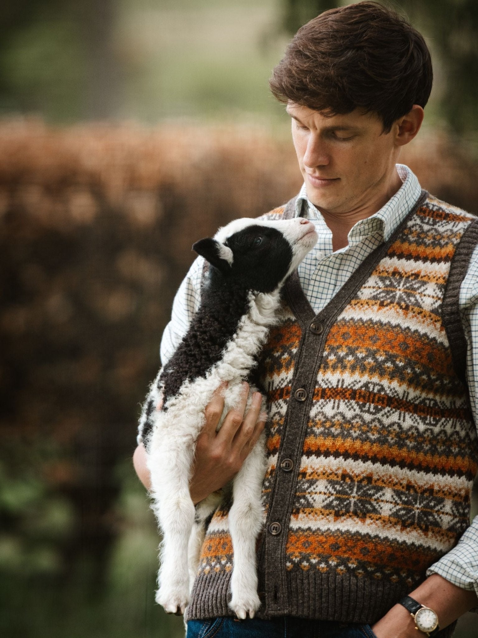 A man in a Campbell's of Beauly Fairisle V-Neck Waistcoat holds a black and white lamb outdoors, gazing down at the animal with affection.