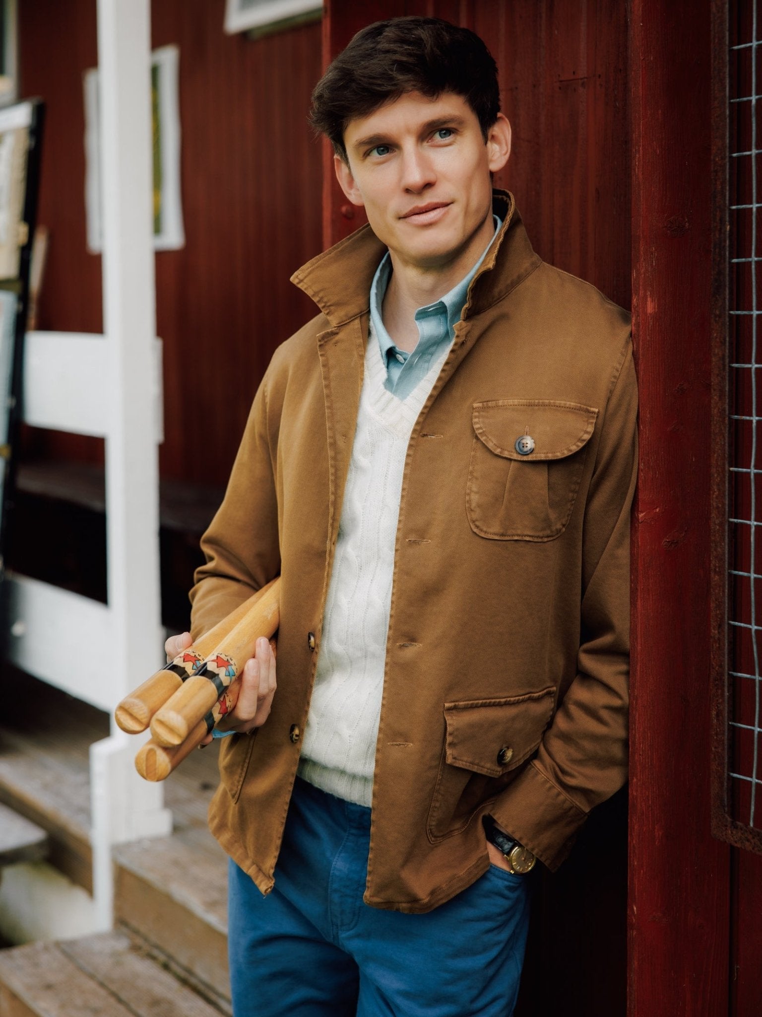 A young man in a Campbell's of Beauly Field Chore Jacket II and blue pants leans against a red wooden wall, holding two wooden rolling pins adorned with colorful bands, gazing thoughtfully into the distance.