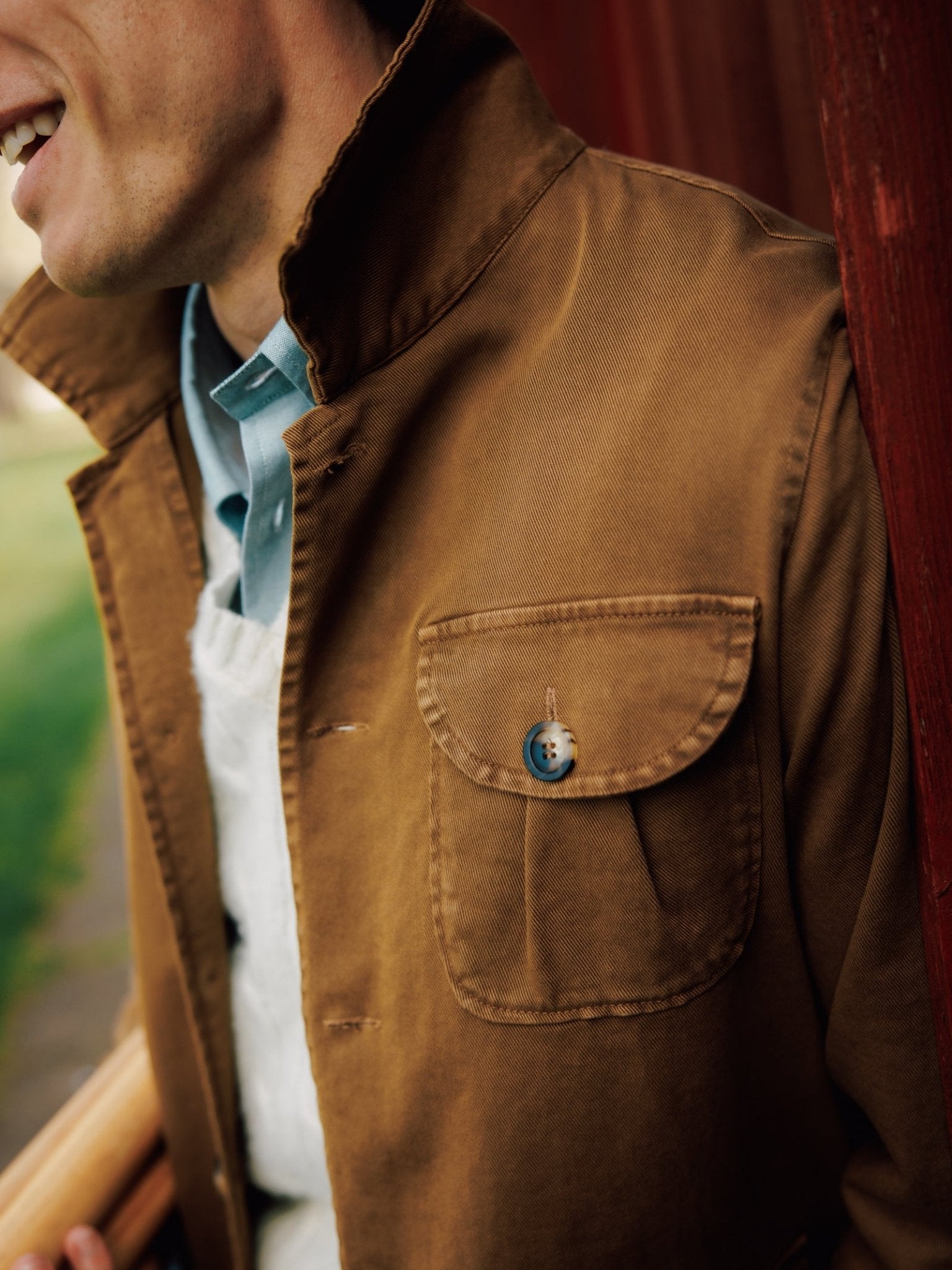 A person smiles near a red wooden surface, wearing Campbell's of Beauly Field Chore Jacket II in brown over a light blue collared shirt and white sweater. The men's cotton jacket features a buttoned chest pocket for added style.