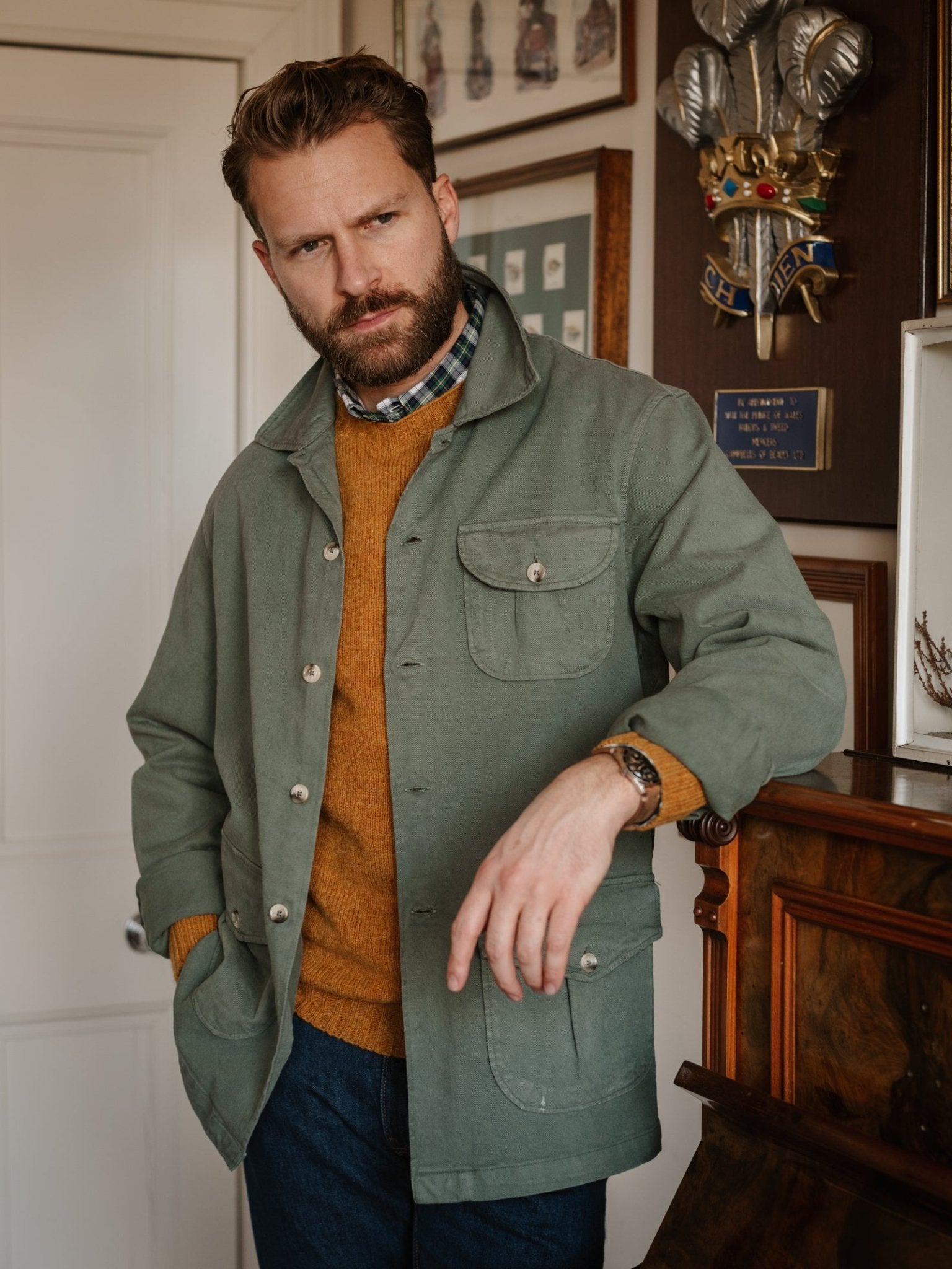 A bearded man with light brown hair stands indoors, wearing a sage green Campbell's of Beauly Field Chore Jacket over a mustard sweater and plaid shirt, leaning on a piano with framed pictures and a coat of arms behind him.