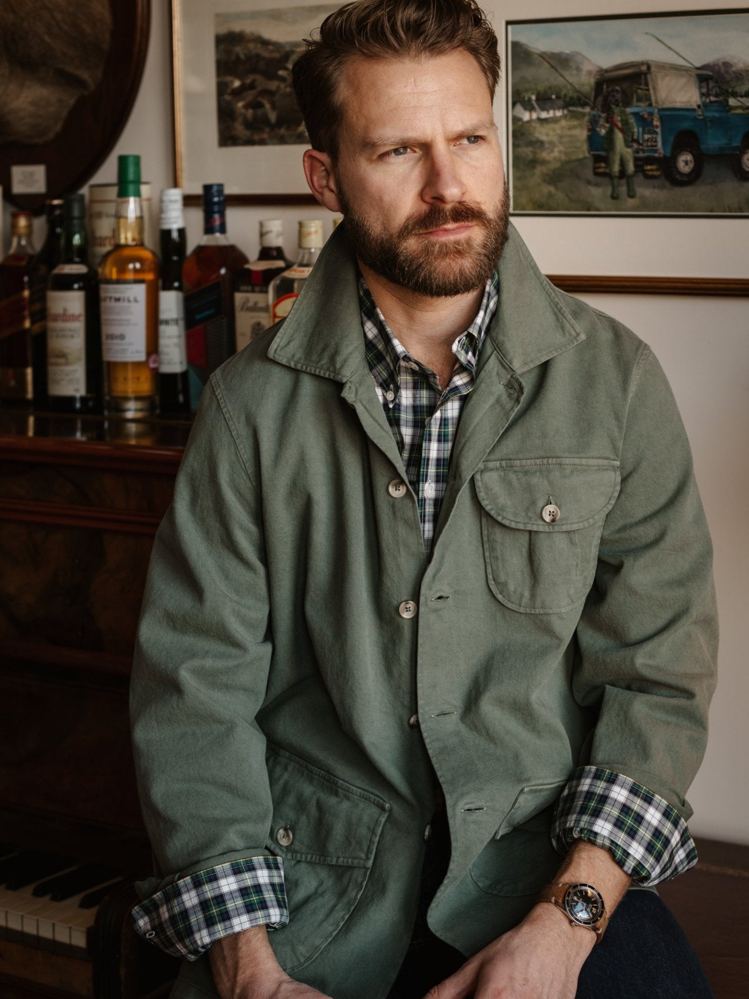 A bearded man with short hair wears a Campbell's of Beauly Field Chore Jacket over a plaid shirt, sitting indoors by a piano and liquor bottles, with framed art visible on the wall behind him.